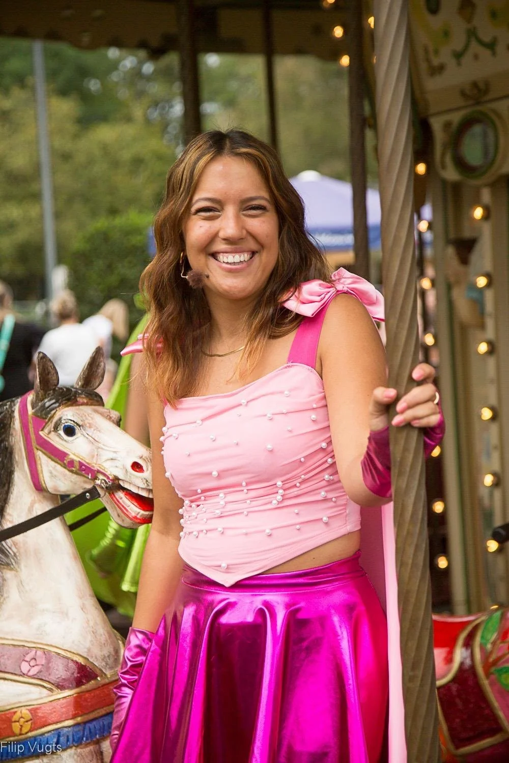 Vrouw glimlachend op een meisje tokkelassa, gekleed in een roze en fuchsia outfit, op een kindervoorstel met een achtergrond van een sprookjesachtig wezen en een groene omgeving.
