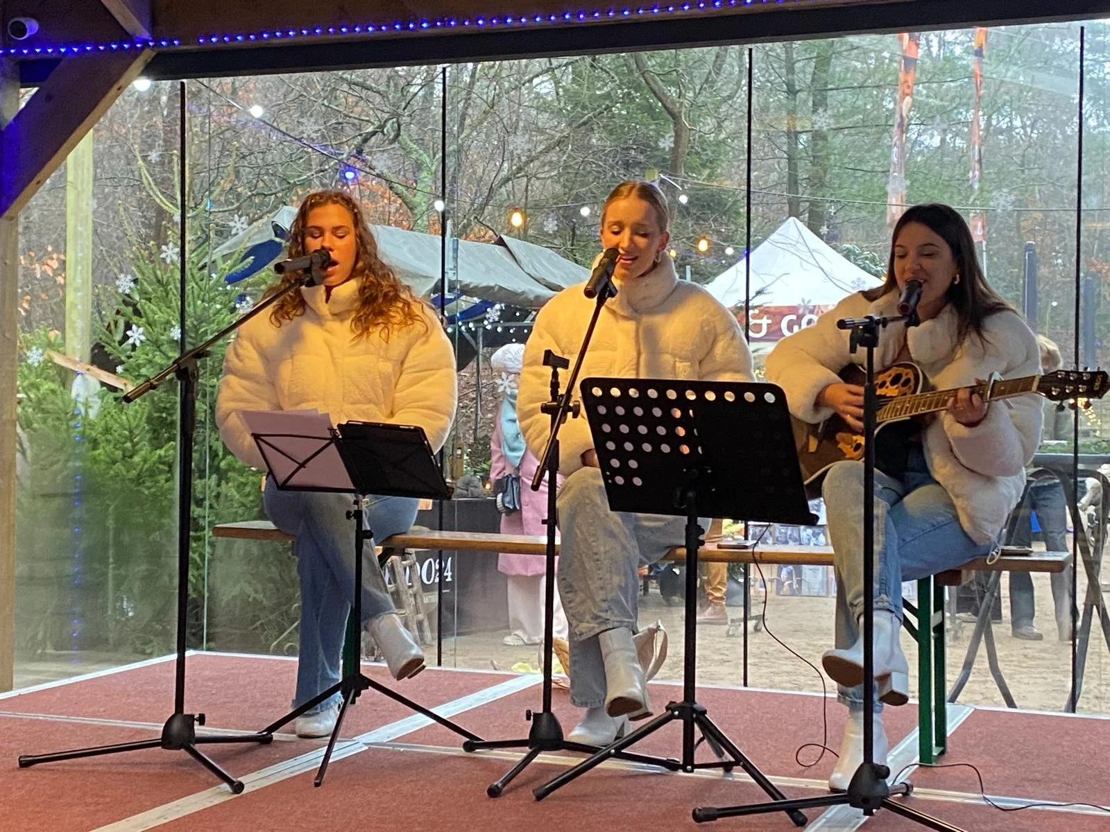 Vrouwelijke muzikanten zingen en spelen gitaar tijdens een live-optreden in een indoorhumoristische setting, met buiten visible bomen en kerstdecoraties.