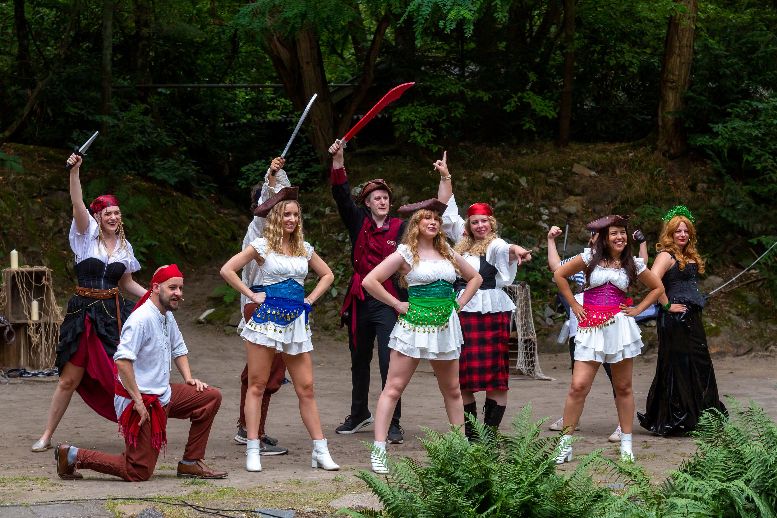 Verschijningsvolle mensen in historische kostuums die dansen en poseren in het bos, met enkele mensen die met beren en piratenachtige hoeden op de achtergrond.