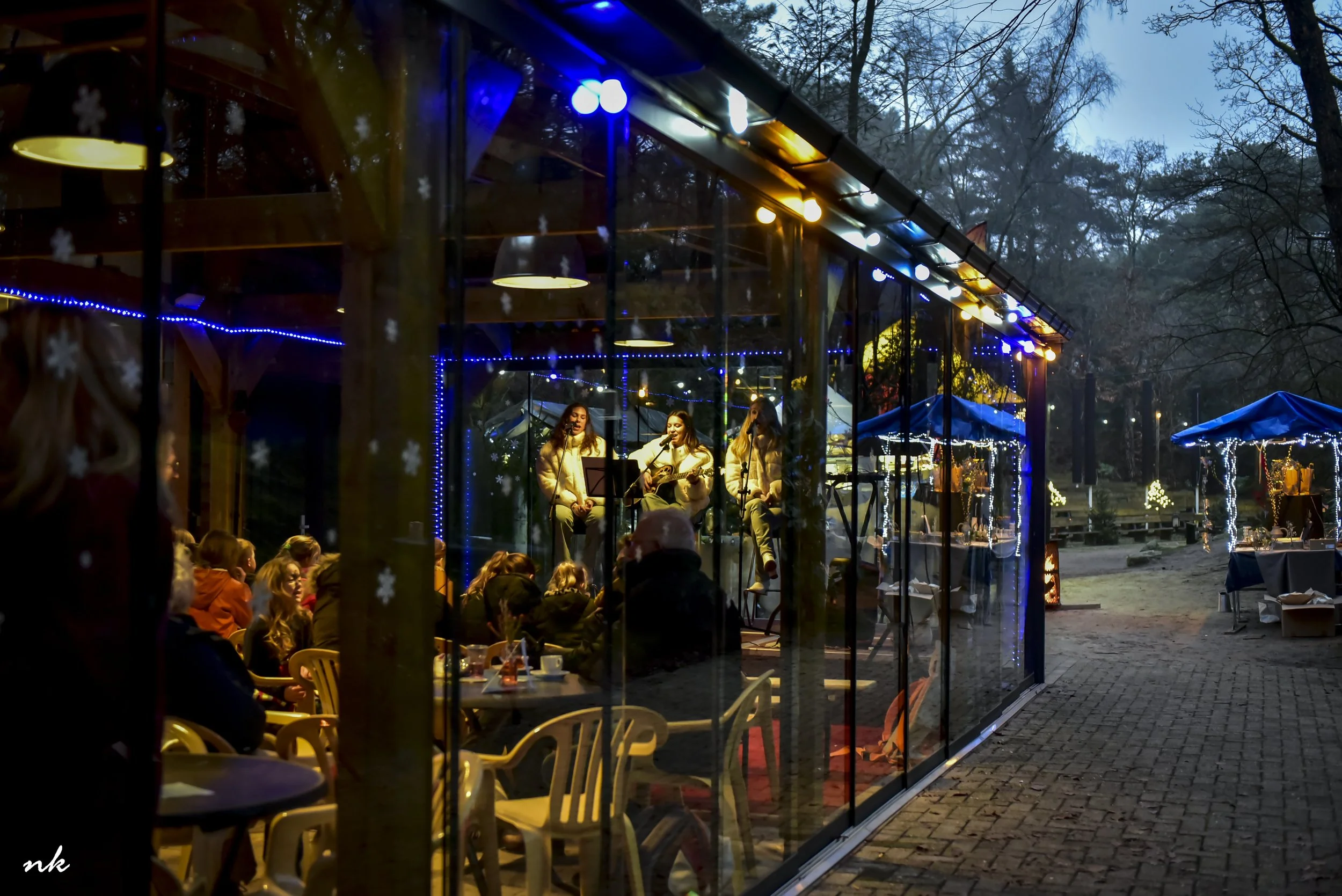 Een glazen serre met feestverlichting en een live muziekoptreden, omgeven door bomen en buiten eetgedeeltes, tijdens de avond.