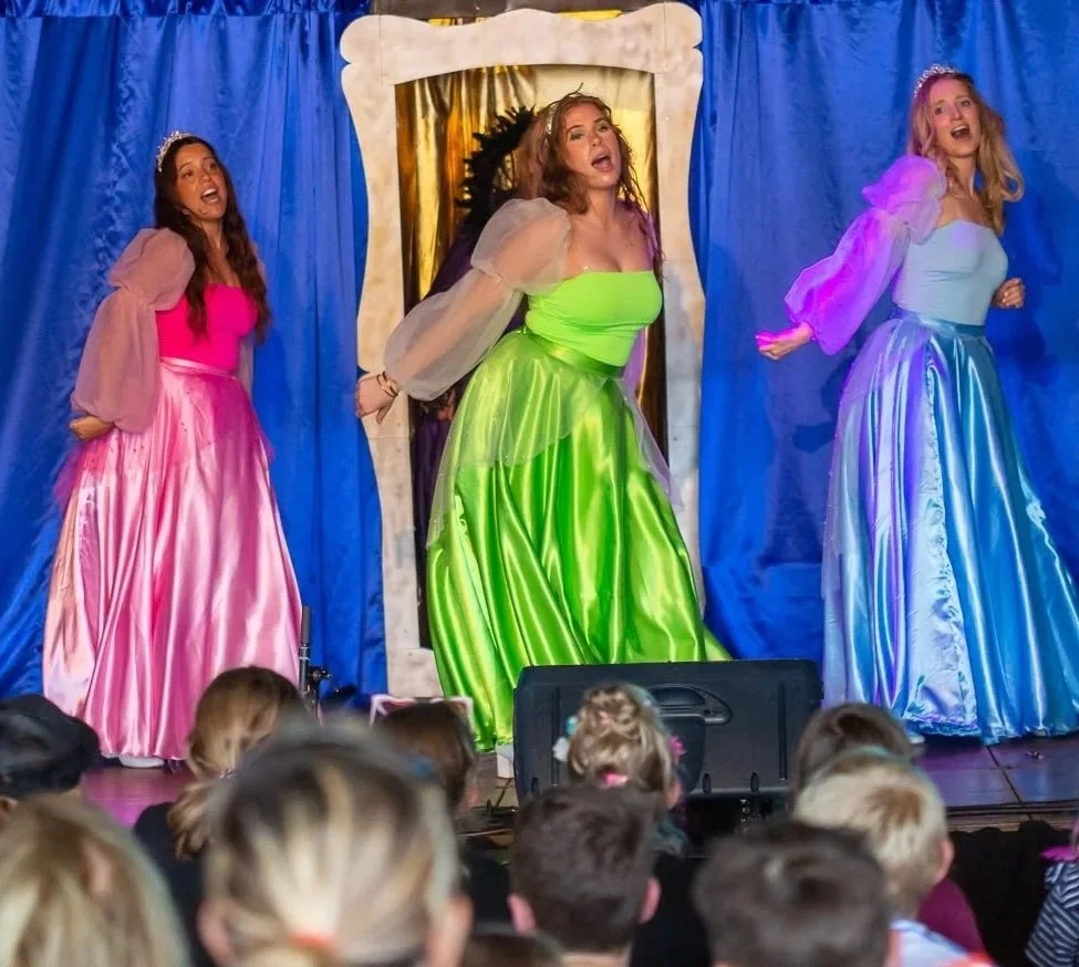 Vier vrouwen in kleurrijke, volledig geplooide jurken die op een podium dansen en zingen, met een publiek van kinderen en volwassenen voor zich.