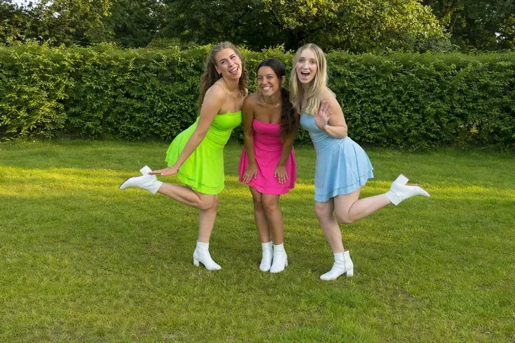 Drie lachende jonge vrouwen in heldere jurkjes en witte laarzen, poseren op een grasveld met groen gebladerte op de achtergrond.