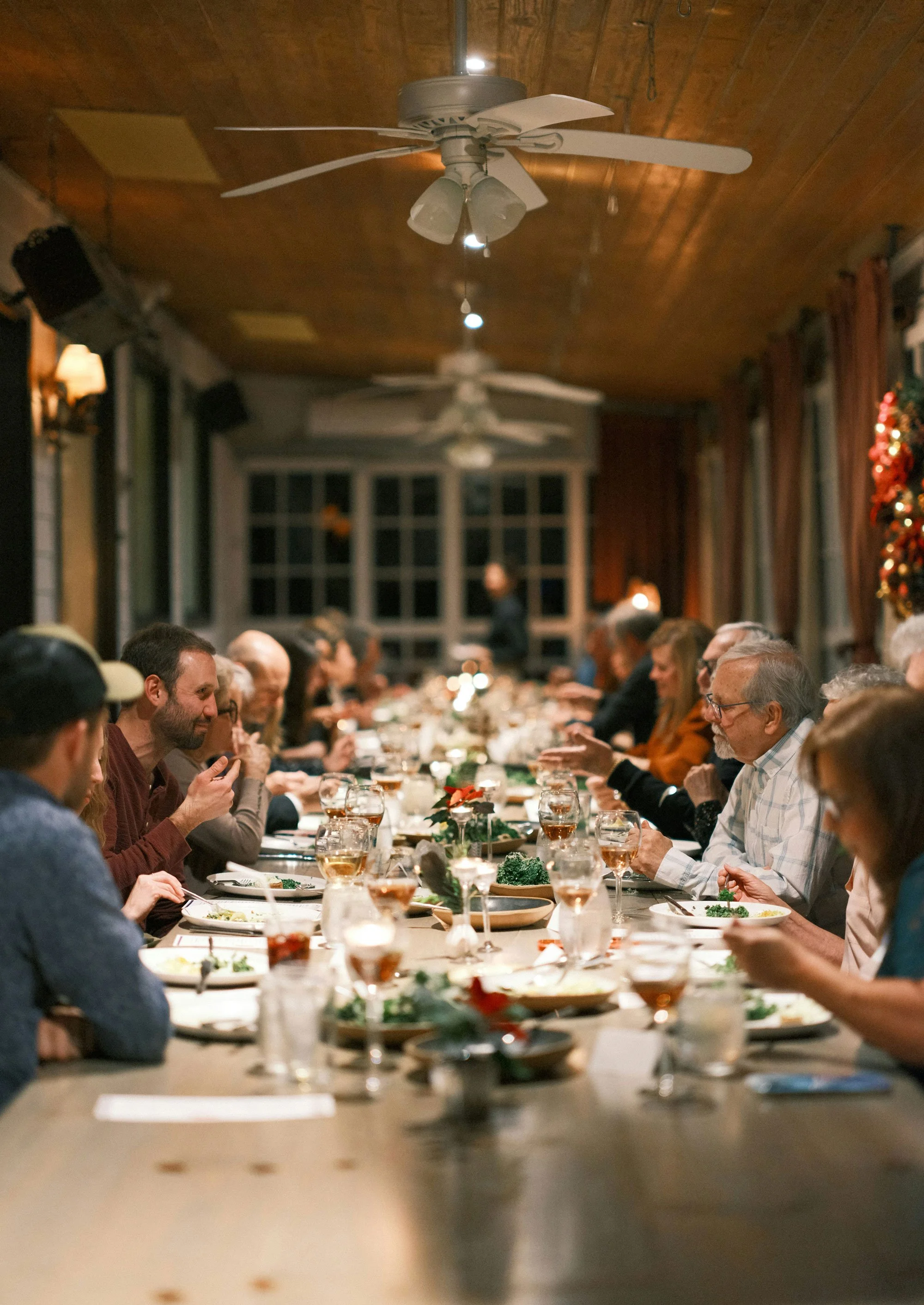 People sitting at a long dinner table enjoying a meal in a warmly lit room with wooden ceiling and ceiling fans during a festive gathering.