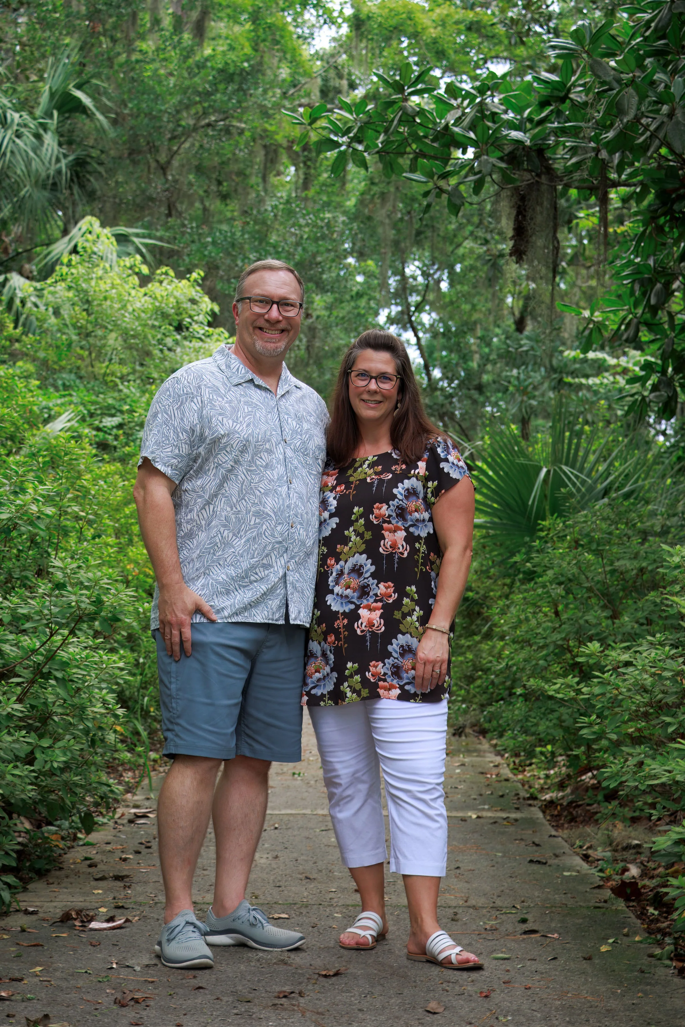 A smiling man and woman standing on a path in a lush, green forest. The man is wearing glasses, a patterned short-sleeve shirt, and shorts. The woman is also wearing glasses, a floral top, and white pants.