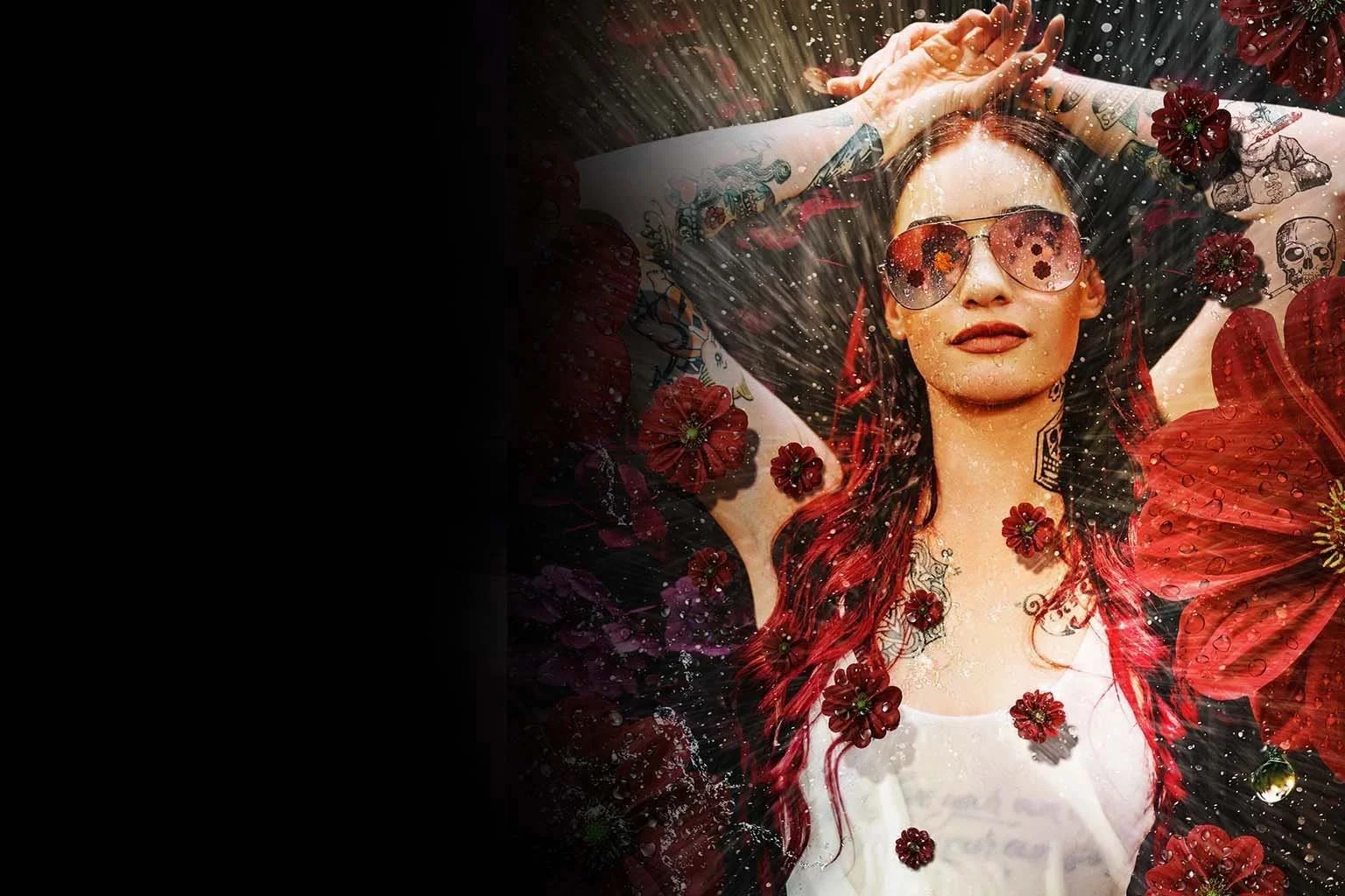 A woman with red hair and tattoos on her arms and neck, wearing sunglasses and a white shirt, is lying on her back with her arms above her head. She is surrounded by red flowers and appears to be under a spray of water or light, creating a sparkling effect.
