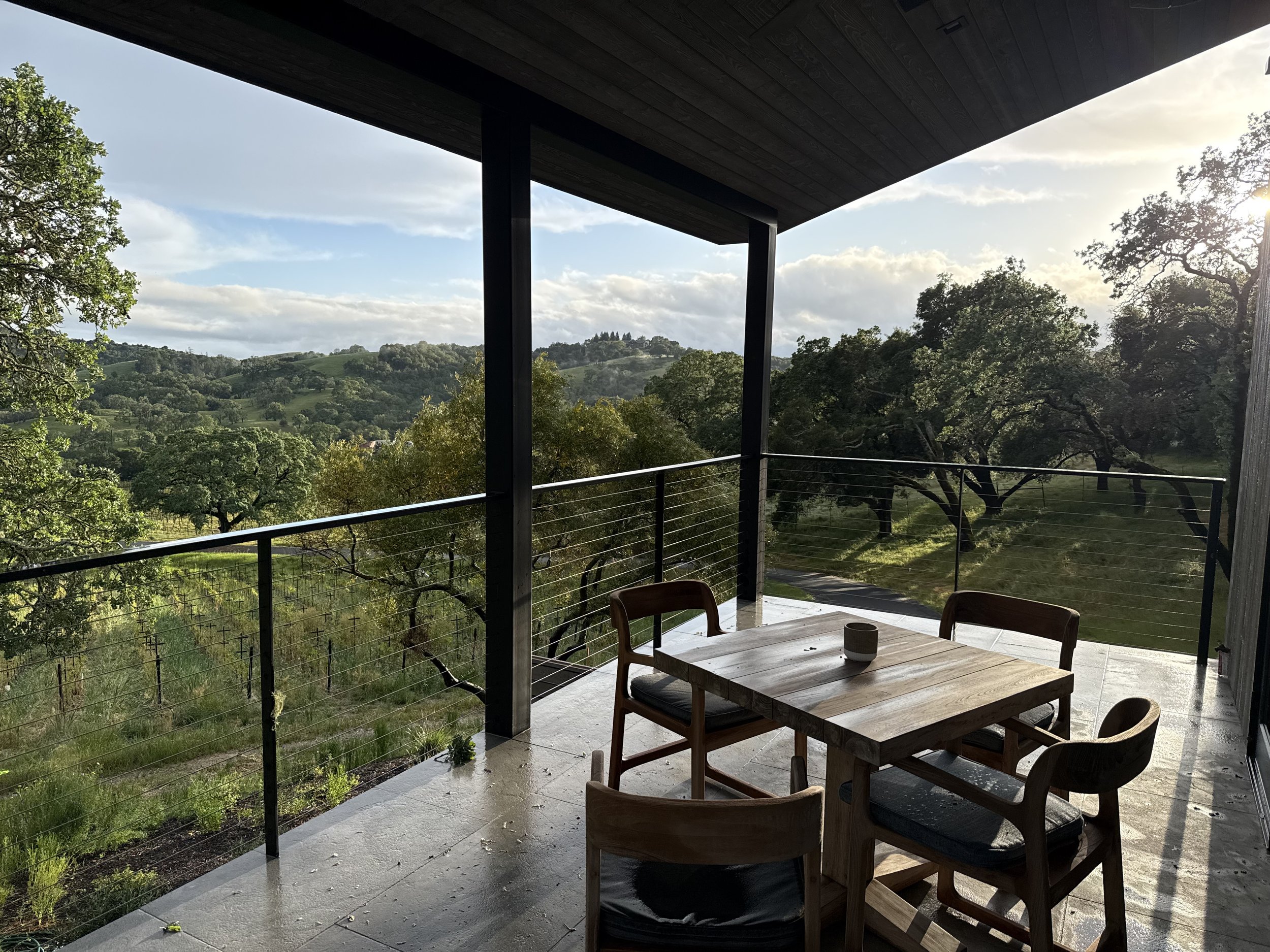 Sunset view from a balcony with a wooden table and chairs, overlooking green rolling hills and trees.