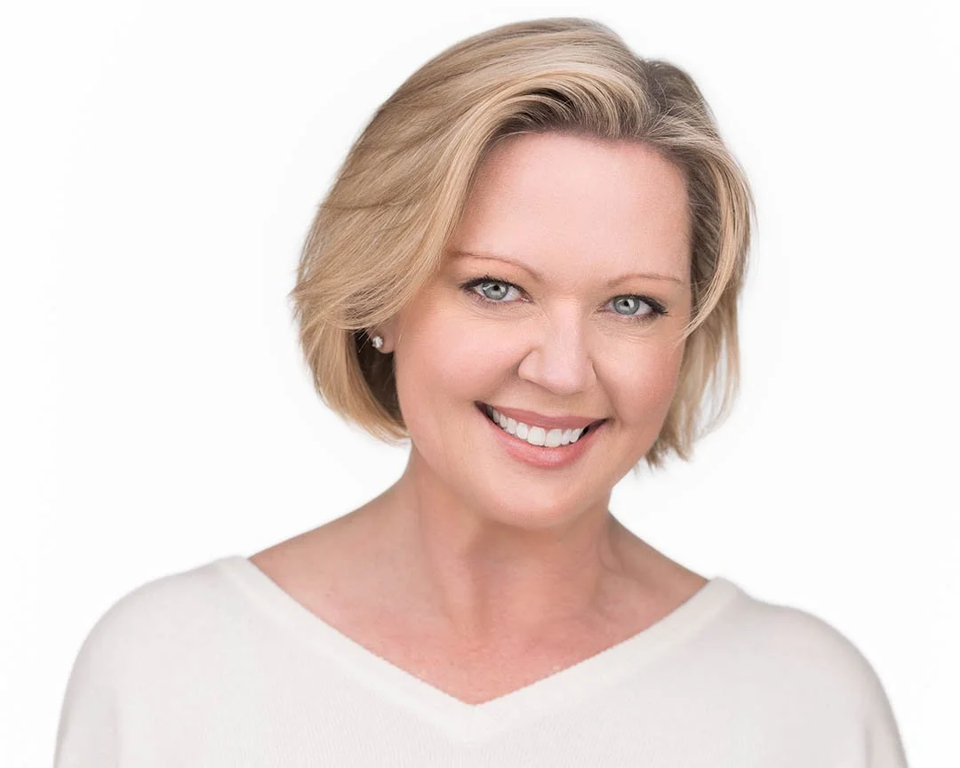 A woman with blonde, shoulder-length hair smiling, wearing a white top and small earrings against a plain white background.