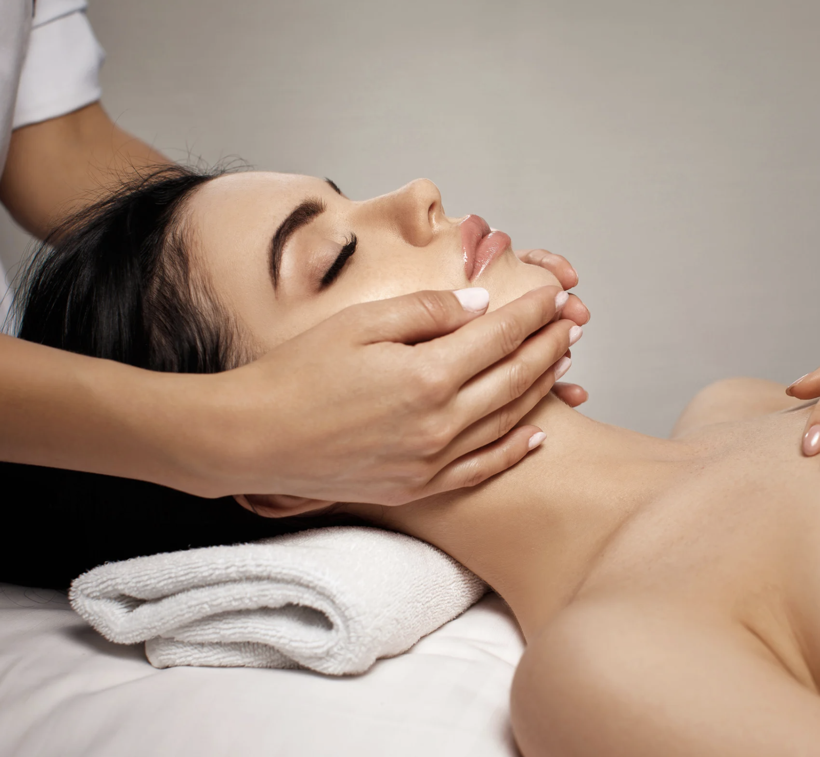 A woman receives a facial massage while lying on a treatment bed with a rolled white towel underneath her head, in a spa or wellness center.