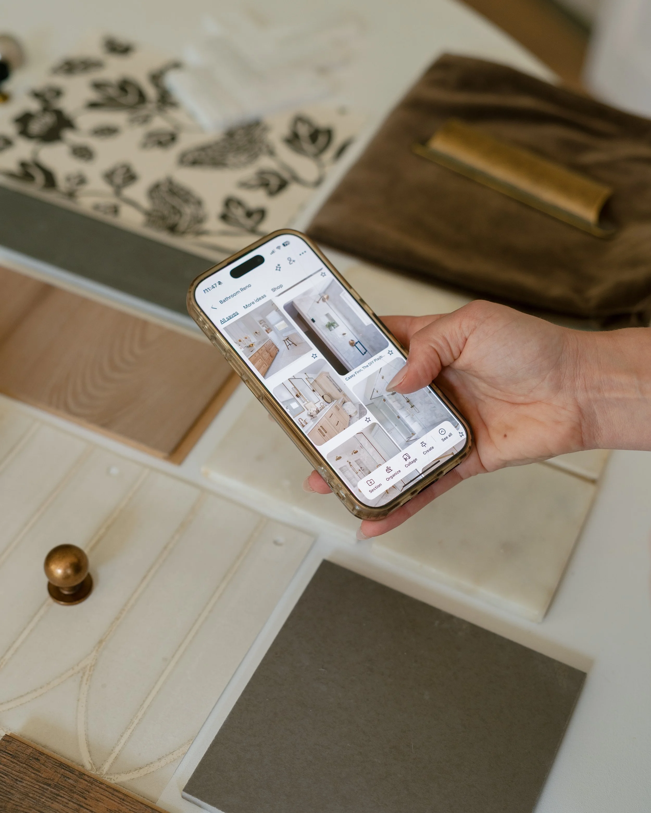 A person holding a smartphone with a home interior design app open, showing images of kitchen and bathroom designs, standing next to a white dresser with a brass-colored handle and a drawer on a hardwood floor.