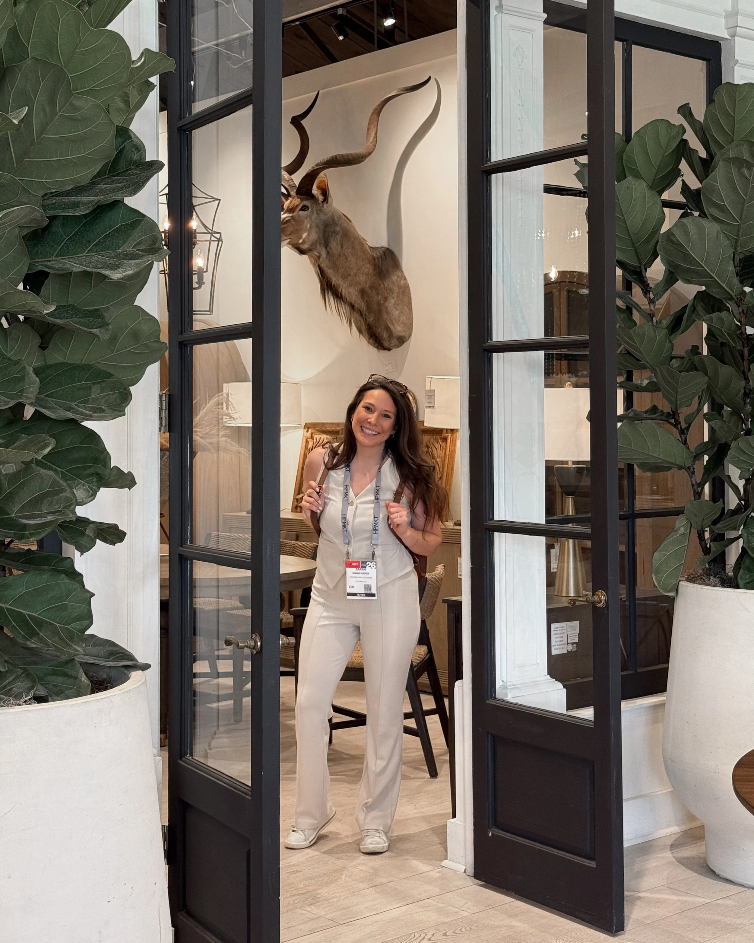 An interior designer at High Point Market in North Carolina. Stands inside Gabby Home's Showroom. Featuring a decorative wall-mounted animal head through a breathtaking black-framed glass door. The room has indoor plants and modern furniture.