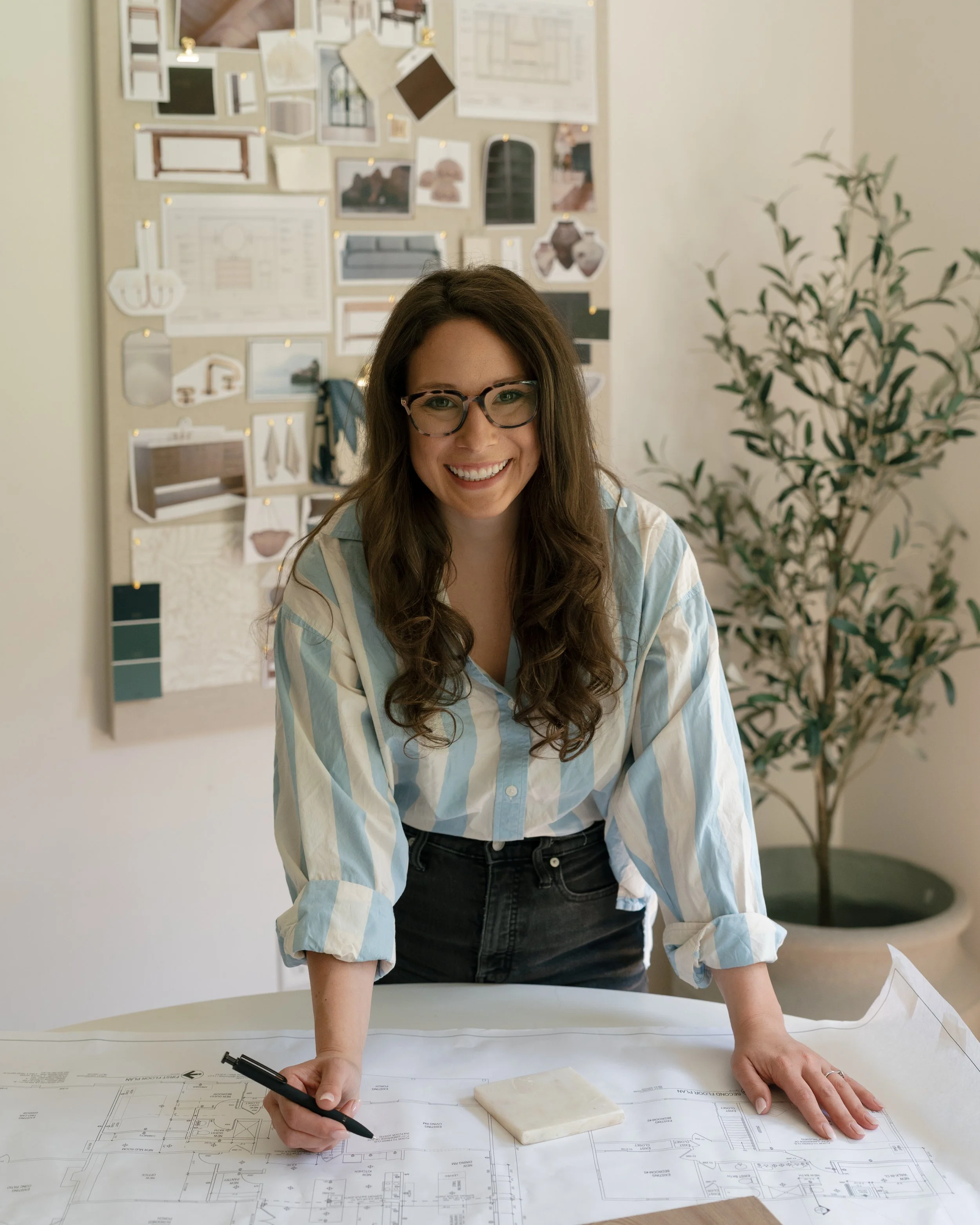 A woman with long brown hair, glasses, and a striped shirt is leaning over a table with her hands on architectural blueprints, holding a black pen in her right hand. There is a small marble block on the blueprints. In the background, there is a corkboard filled with various photos, sketches, and notes, and a potted plant to the right.