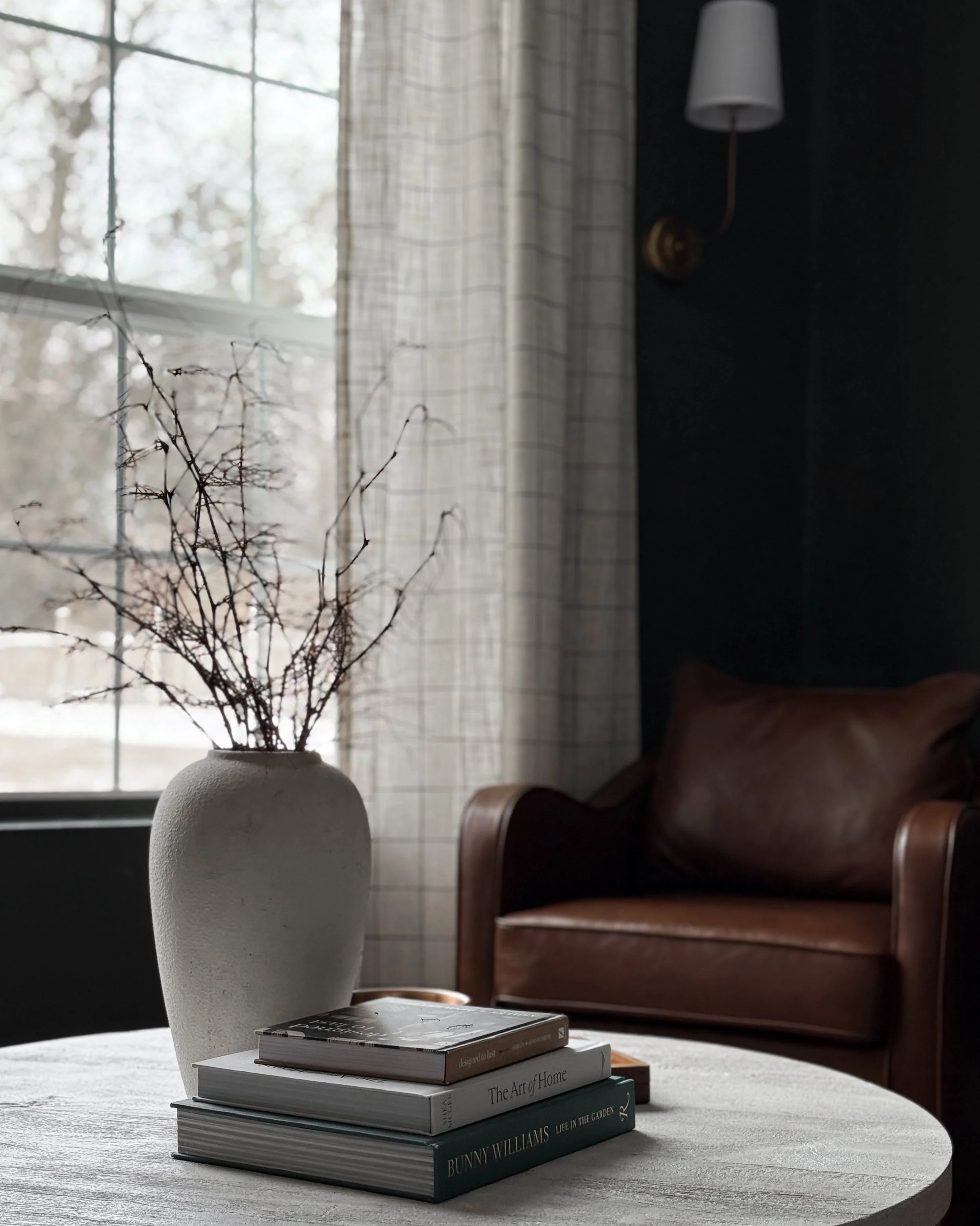 Moody Bar Room Design. Featuring a thrifted vase with dried stems on a coffee table. Dark green (SW Shade Grown 6188 walls). Plaid curtains, and a brown leather chair with gold sconce on the wall.