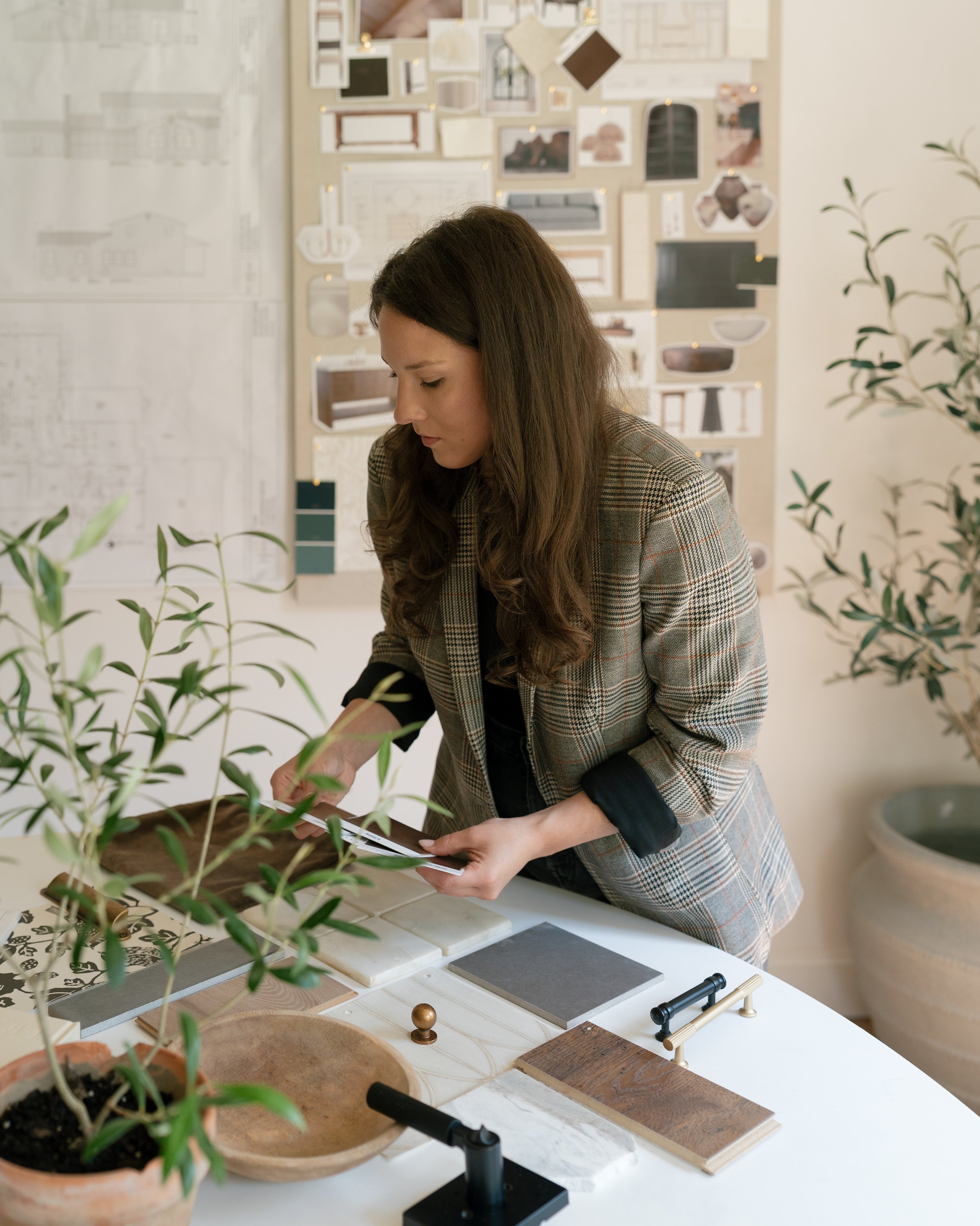 Woman examining design or material samples at a table in a design studio, with greenery and design sketches on the wall behind her.