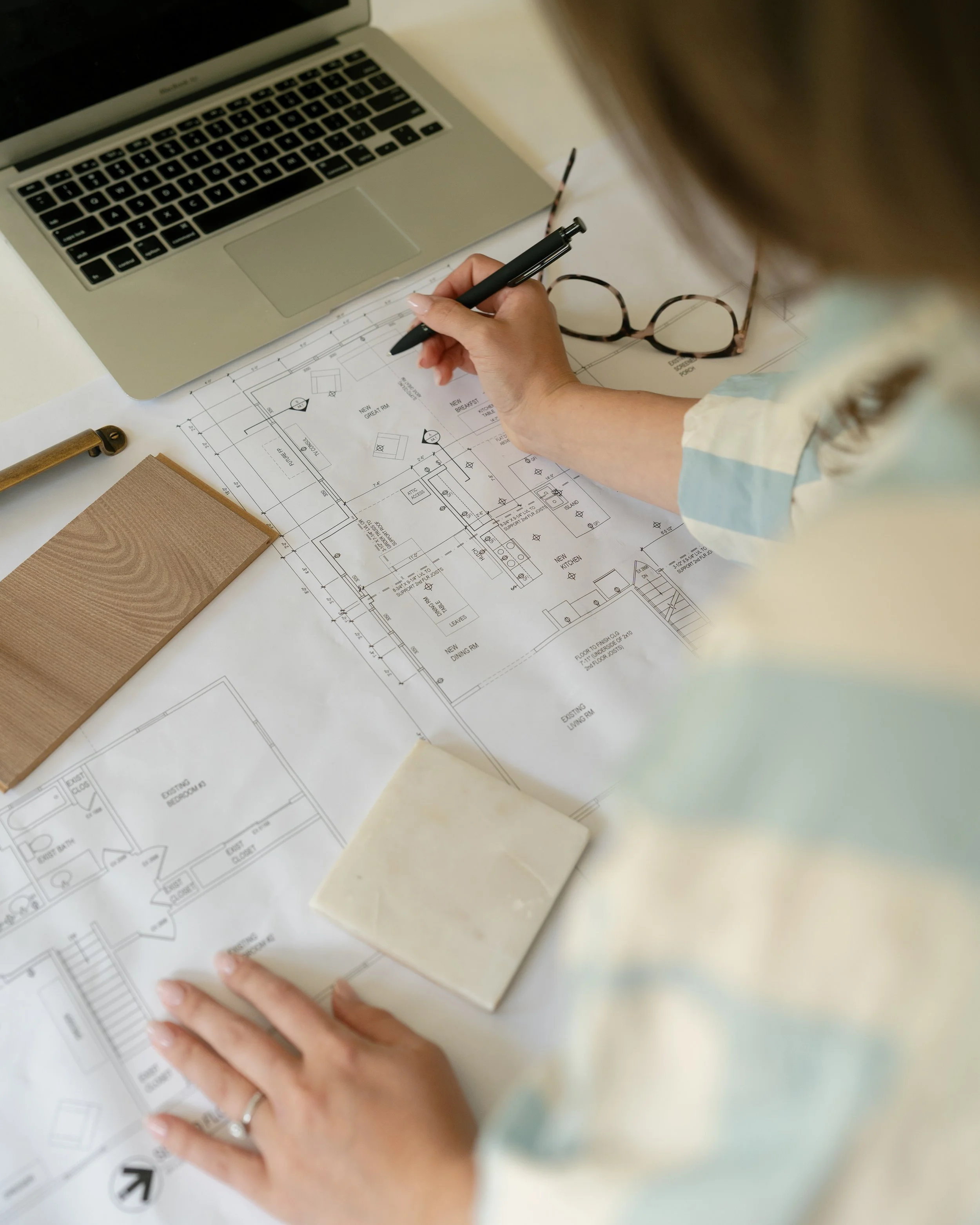 An interior designer (Sarah Sherer) reviewing architectural blueprints on a white table, with an MacBook Air laptop, eyeglasses, a brick-colored notebook, and a small white marble cube nearby.