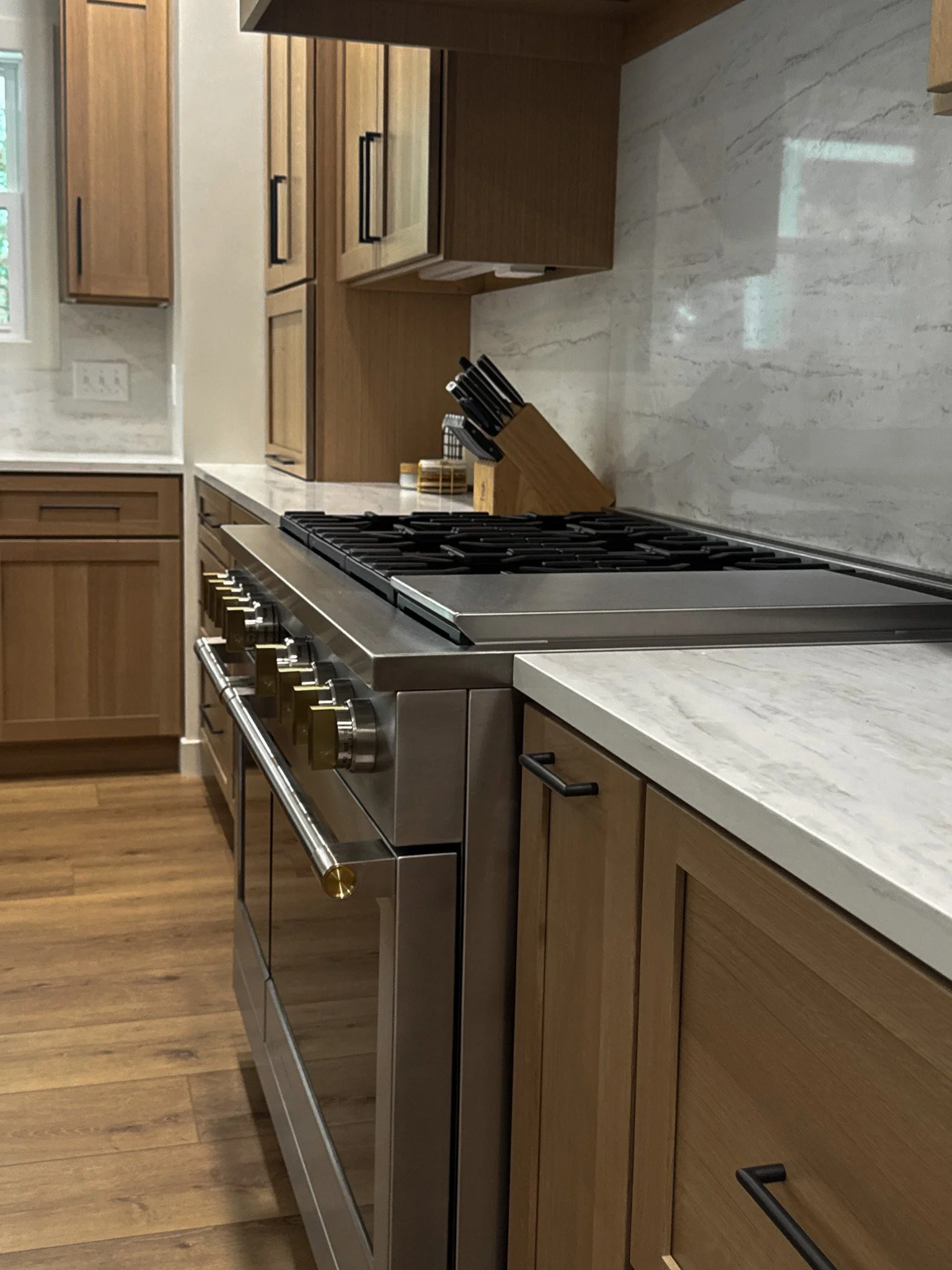 A modern kitchen with wooden cabinets, a quartz countertop, a gas Monogram stove, and a knife block with knives. There is a window with white trim and an electrical outlet on the wall.