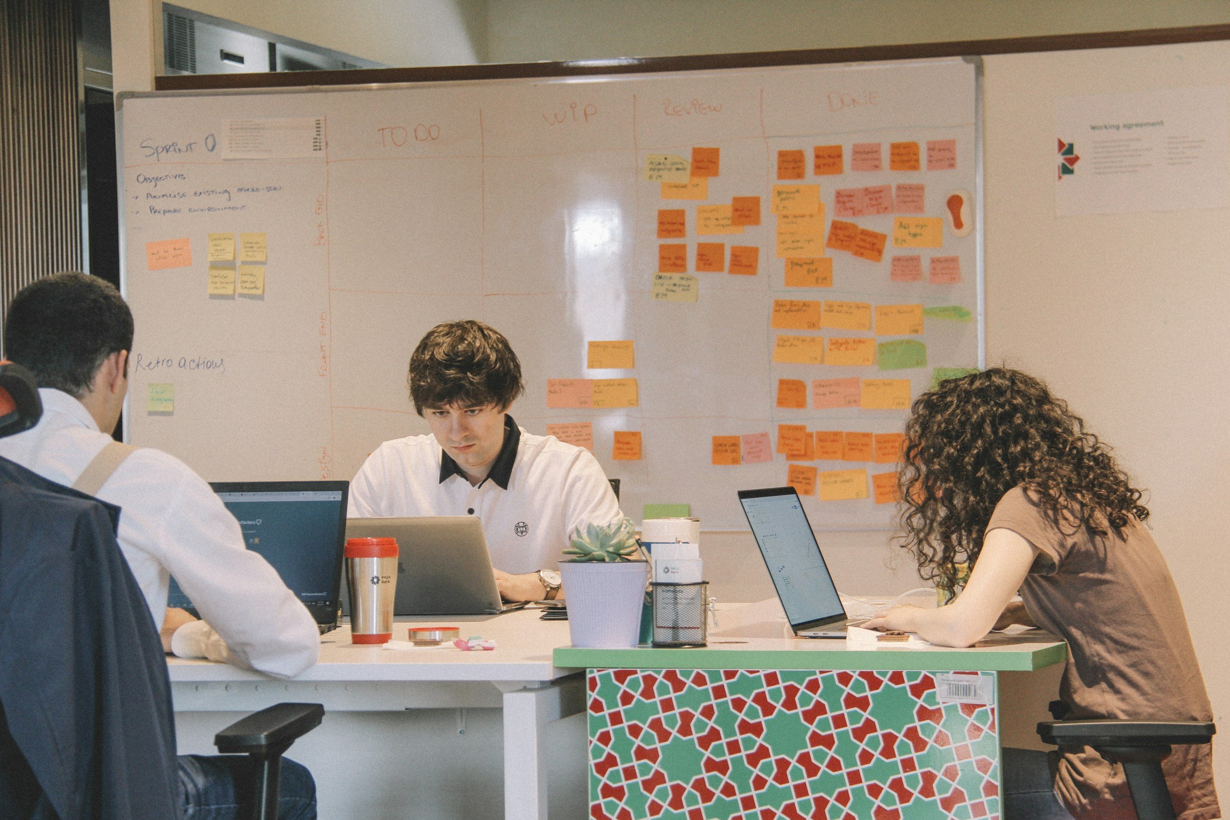 A diverse group of people working at a table with laptops and notebooks in a collaborative office environment. There is a large whiteboard behind them filled with sticky notes and handwritten notes for a project.