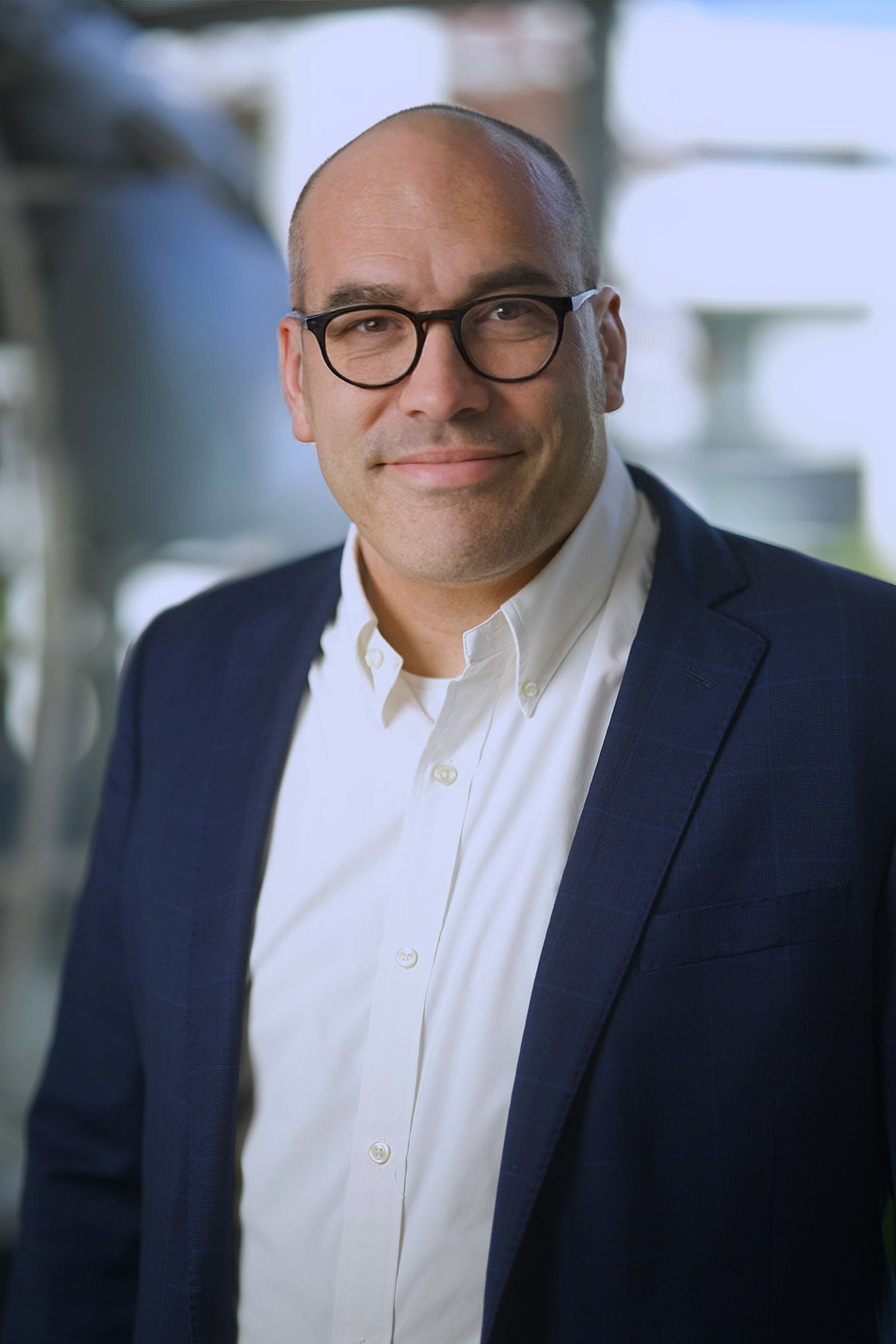 A portrait of a man wearing a white shirt, dark blazer, and black glasses, smiling in a bright indoor setting.