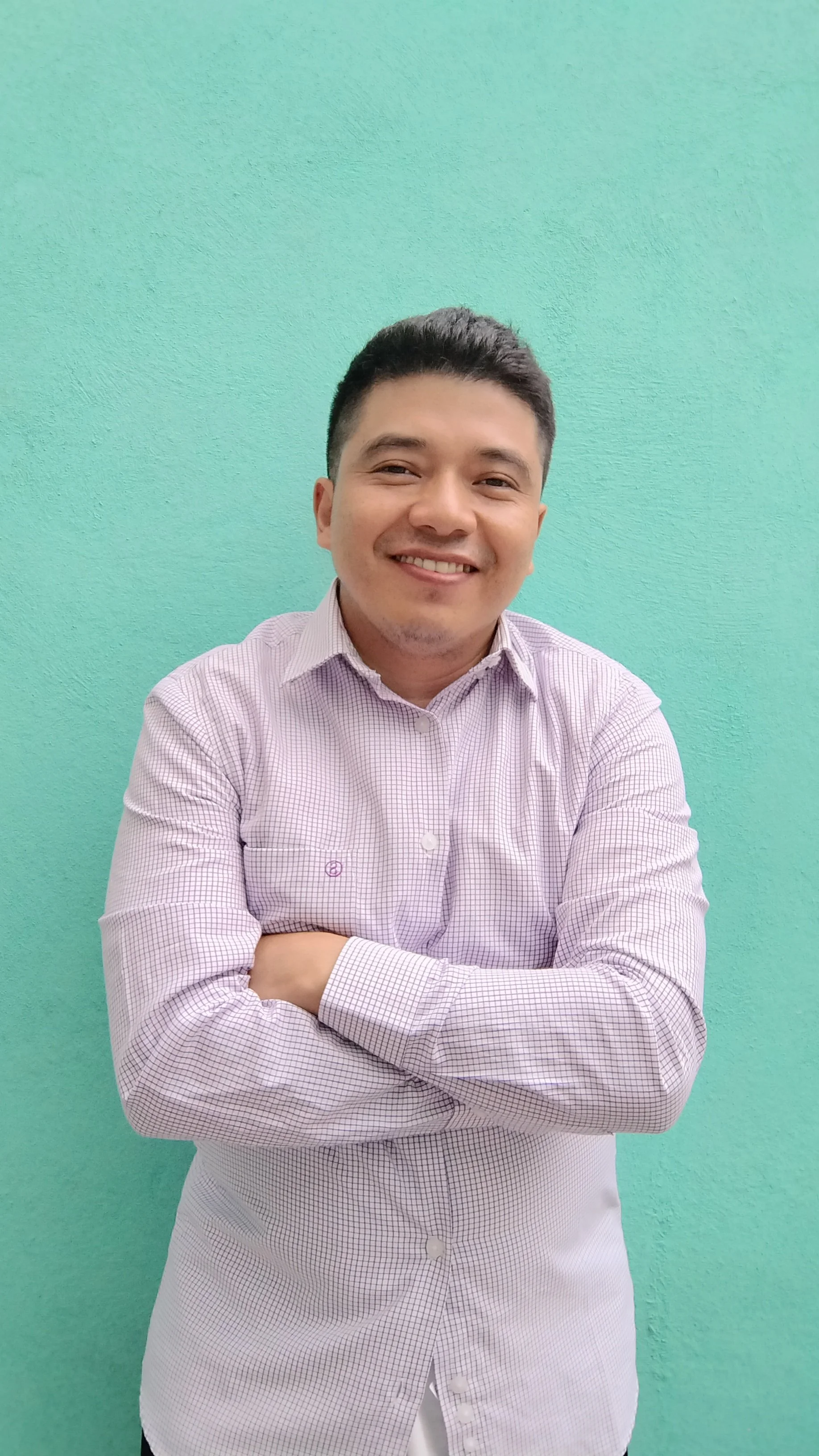 Un hombre joven con cabello oscuro y piel clara, sonriendo y con los brazos cruzados, lleva puesta una camisa de vestir con cuadros pequeños en tonos rosados y blancos, y está de pie frente a una pared de color turquesa.