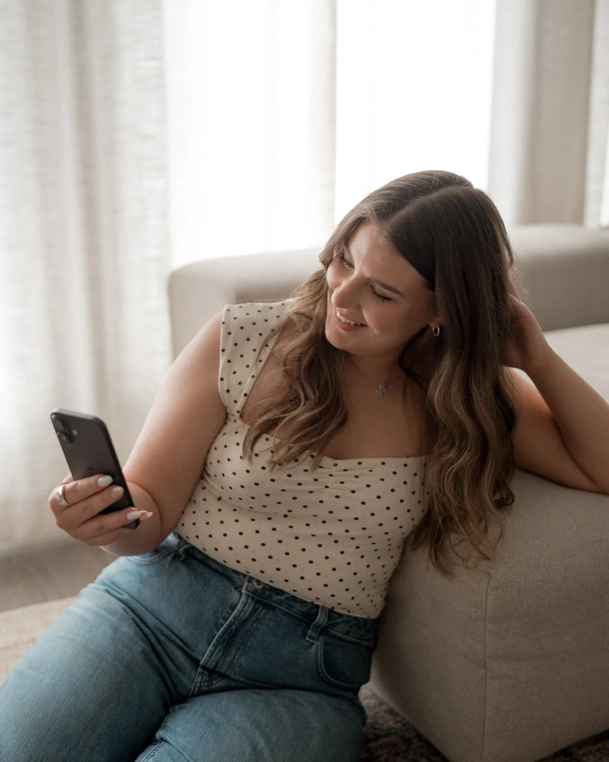 A woman with long wavy hair sitting on a sofa, looking at her phone and smiling.