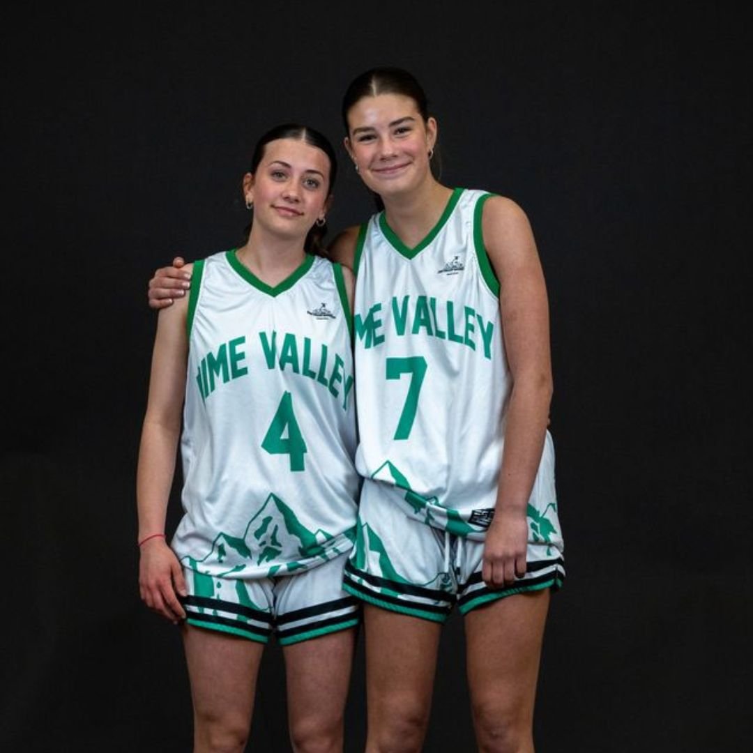 Two young female basketball players standing together in team uniforms against a black background, with one player placing her hand on the other's shoulder. They are smiling.