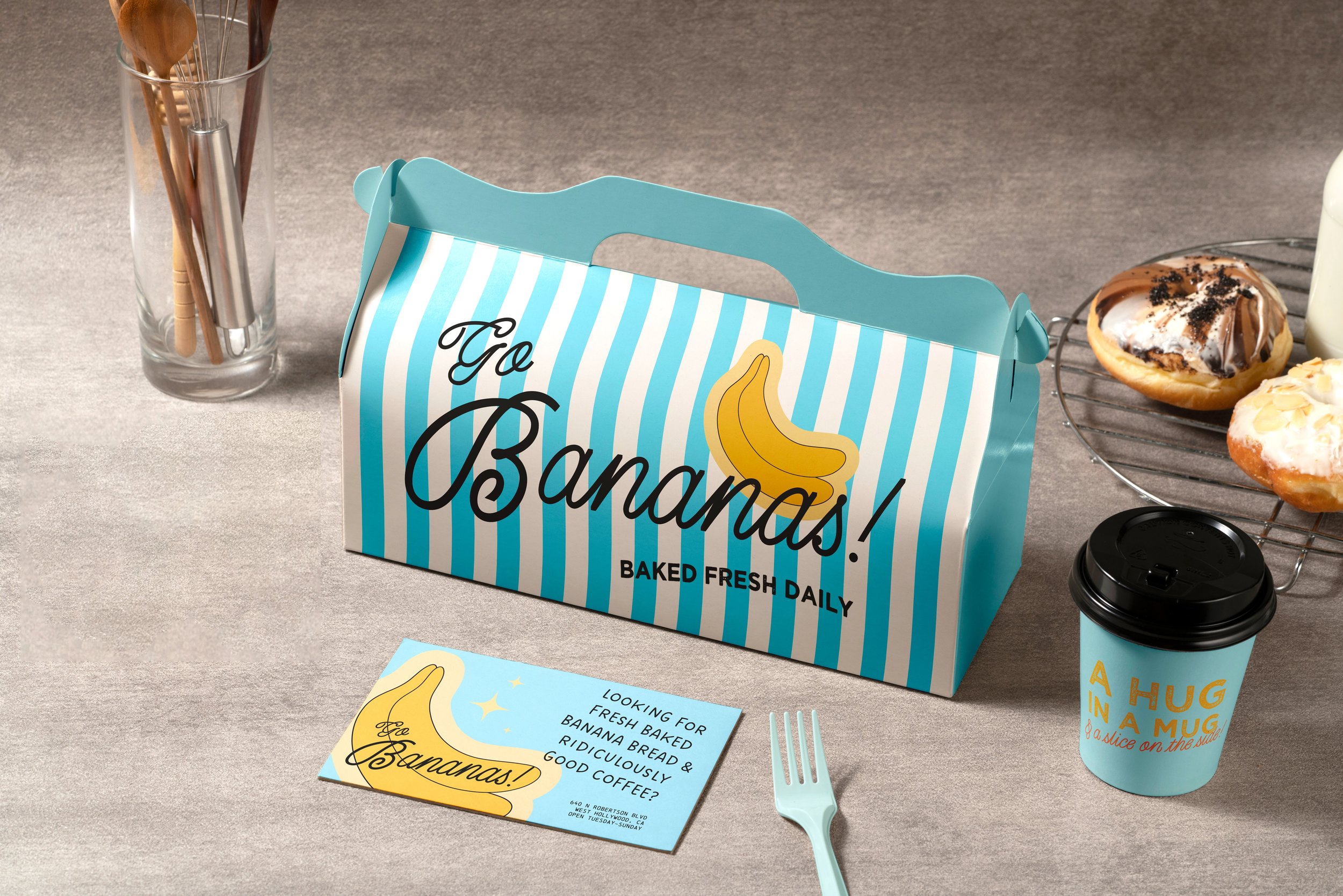 A blue striped box with 'Go Bananas! Baked Fresh Daily' written on it, a cup with a lid that says 'A hug in a mug & a slice on the side,' two donuts on a cooling rack, a card with banana illustrations, a fork, and a glass with utensils inside on a gray surface.