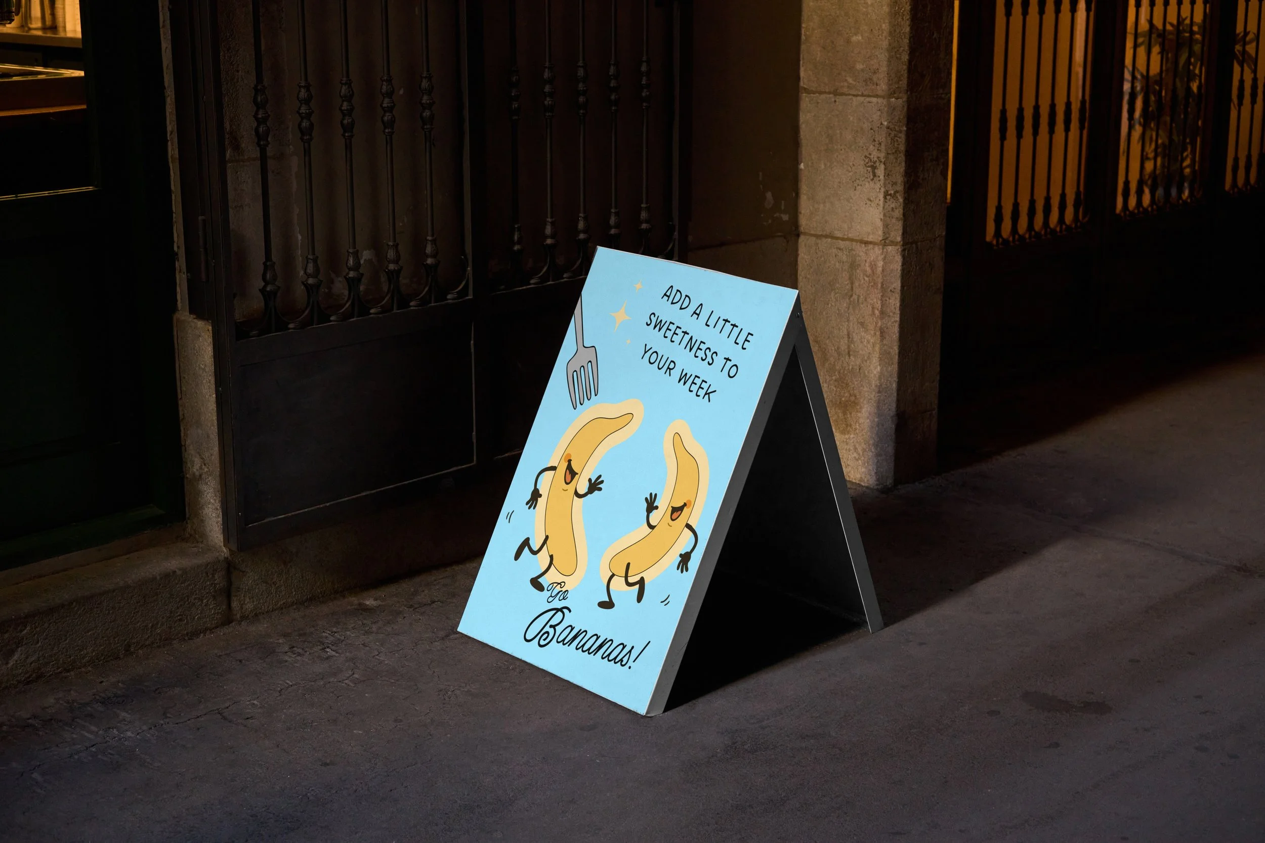 A blue A-frame sidewalk sign with animated bananas on it, depicting two bananas with smiling faces and arms, walking and running, accompanied by stars and a fork. The sign reads 'Add a little sweetness to your week, ya bananas!' and is placed on a sidewalk outside a building with a black gate and stone wall.