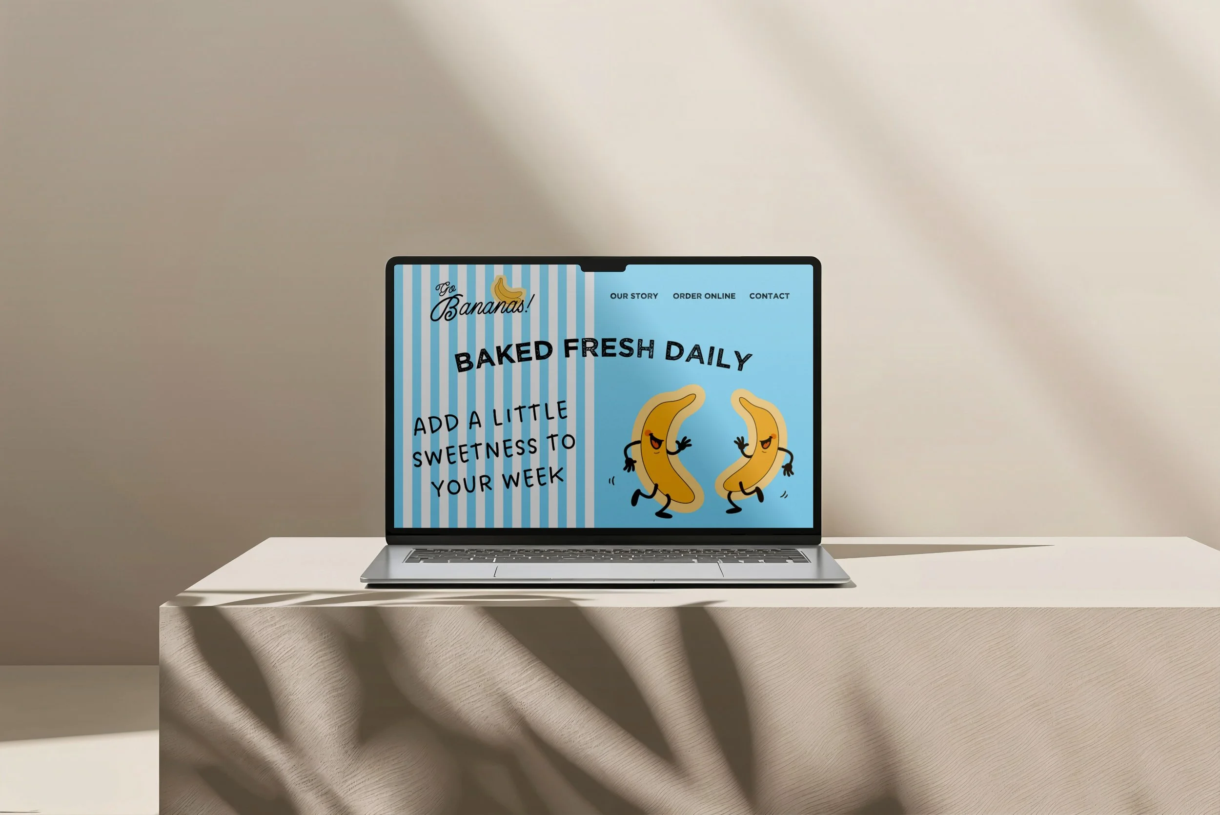 Laptop with a website for a bakery called Go Bananas, showing animated bananas and text about baked fresh daily and adding sweetness to your week, on a white table with shadows and a plain beige wall background.