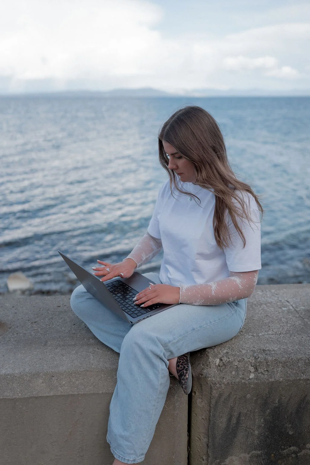 A young woman sitting on a concrete ledge by the water, working on a laptop, with an overcast sky in the background.