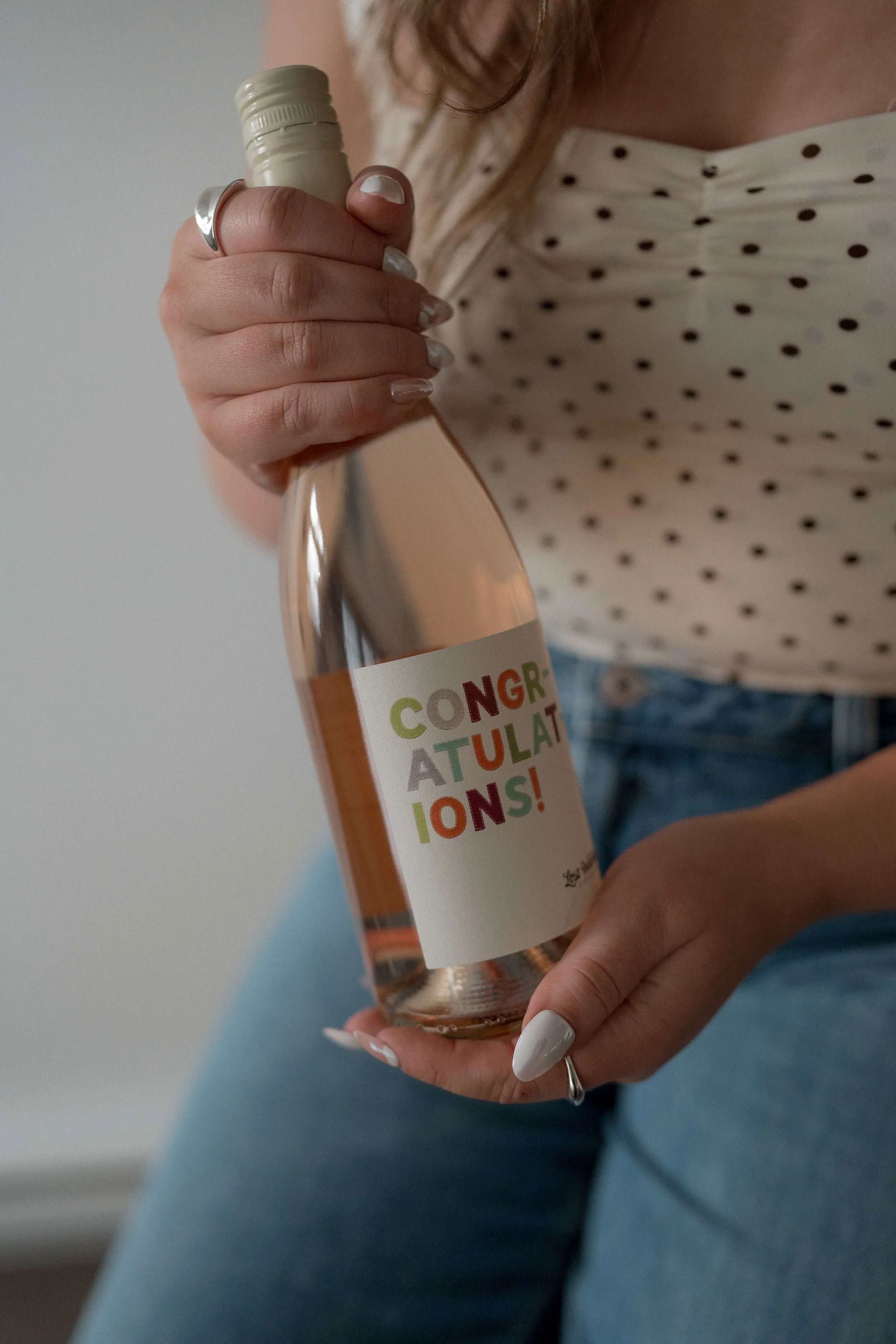 Person holding a small glass bottle with a colorful celebratory label that reads 'CONGRATULATIONS!' in multicolored letters, wearing a white polka dot shirt and blue jeans.