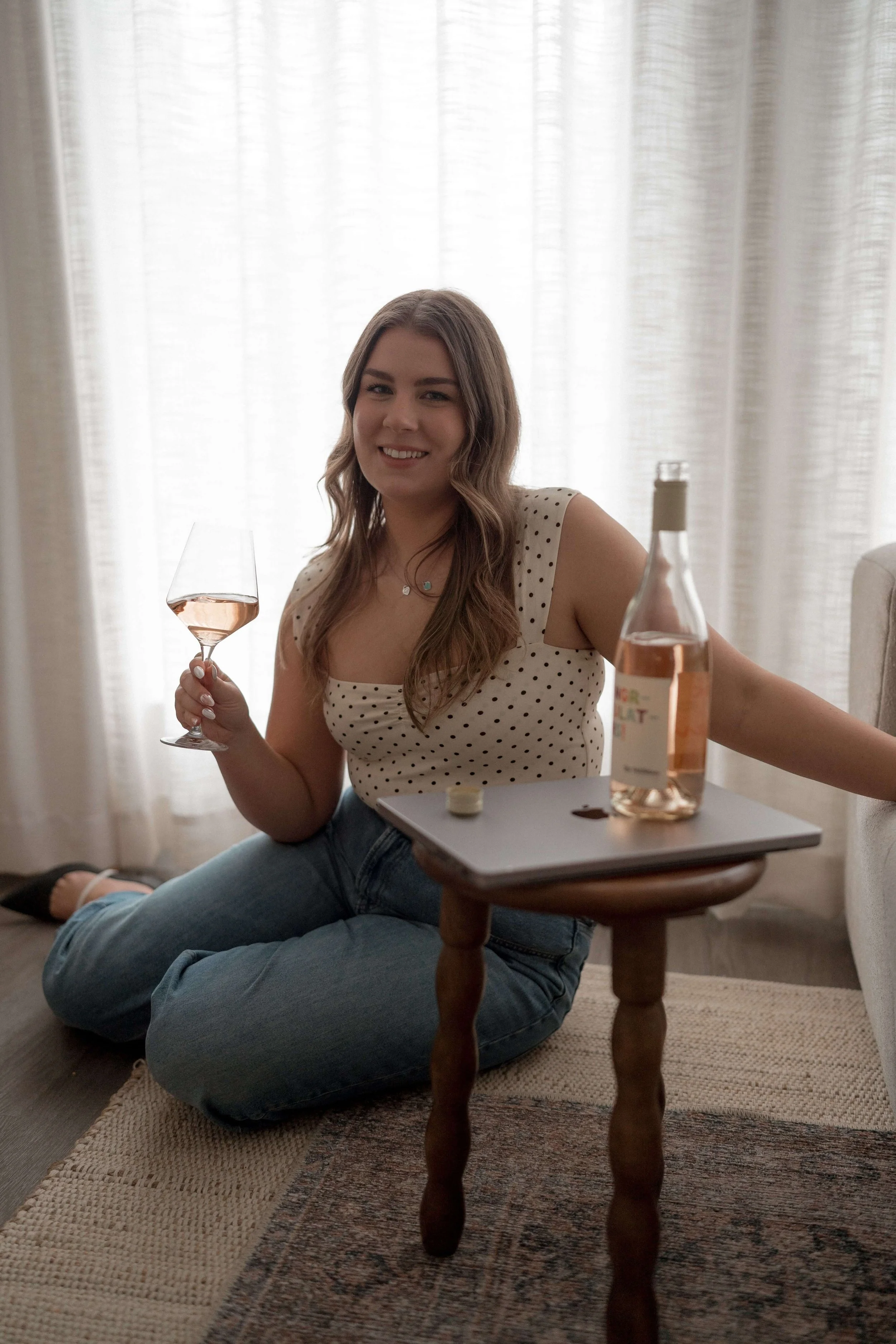 A woman sitting cross-legged on a rug in a living room, smiling and holding a glass of rose wine, with a bottle of rosé wine and a laptop on a small table in front of her. She has long, wavy brown hair and is wearing a white polka-dot sleeveless top and jeans.