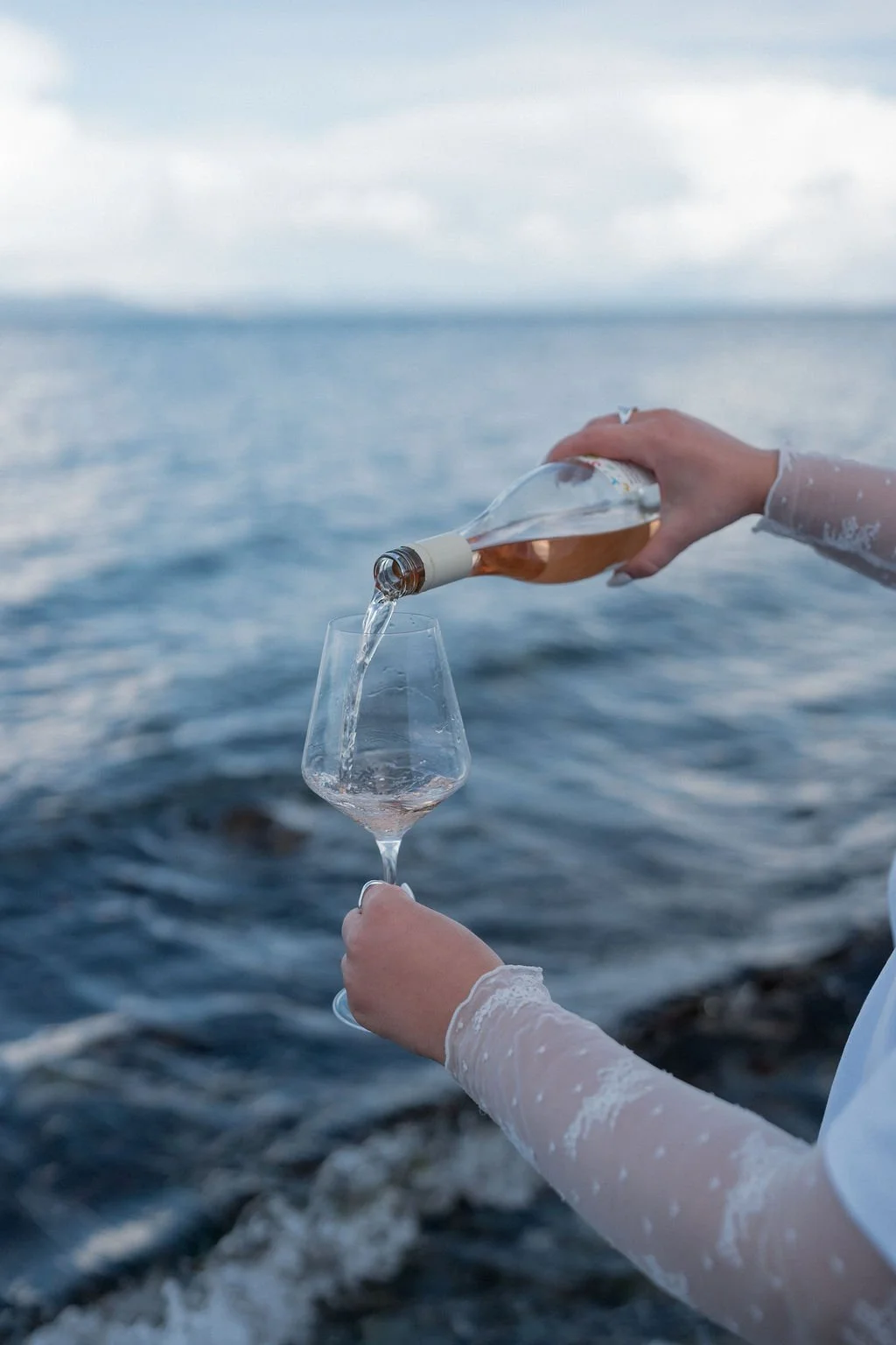 A person in a long-sleeved white dress pours rosé wine from a bottle into a wine glass against a backdrop of the ocean and cloudy sky.