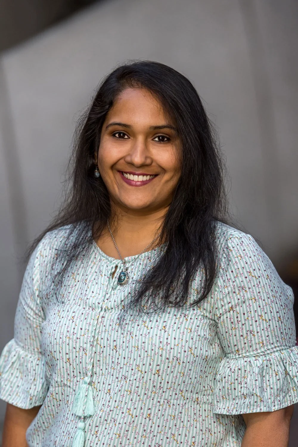 A woman with long black hair, smiling, wearing a light-colored, striped blouse with a necklace and earrings, standing against a neutral background.