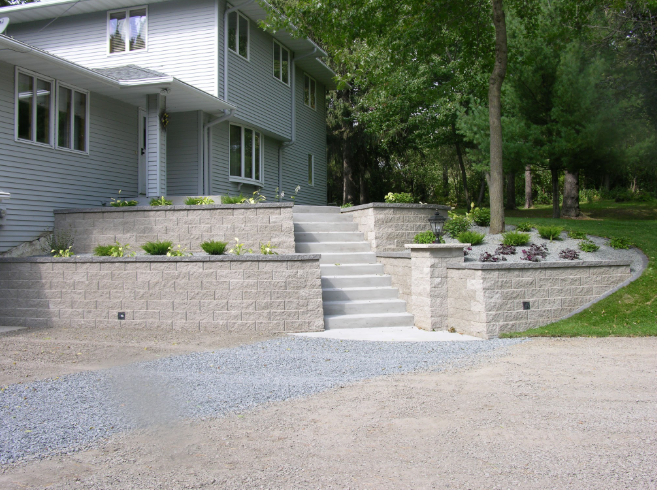 Stamped concrete stairs leading up to a multi-level landscaped garden with stone retaining walls, plants, and trees in front of a light gray house.