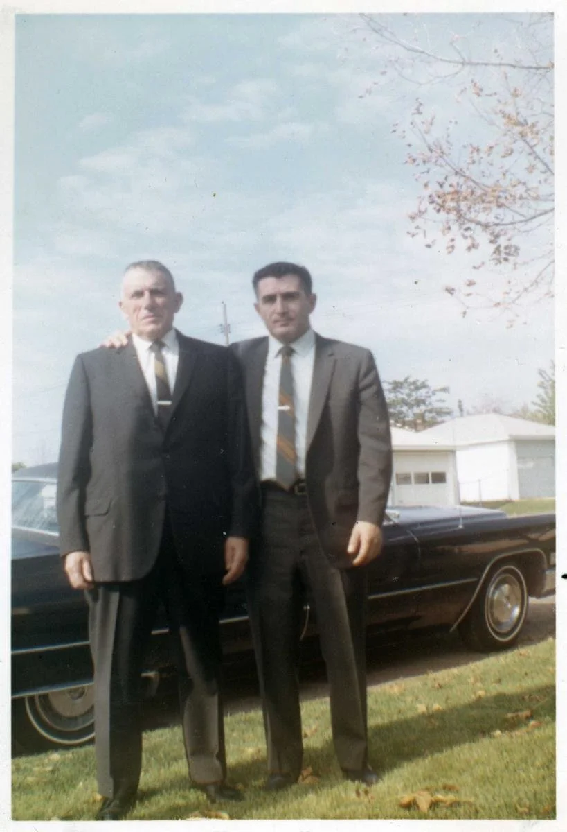 Two men dressed in suits standing outdoors on grass, with a black car behind them and a house in the background, under a partly cloudy sky.