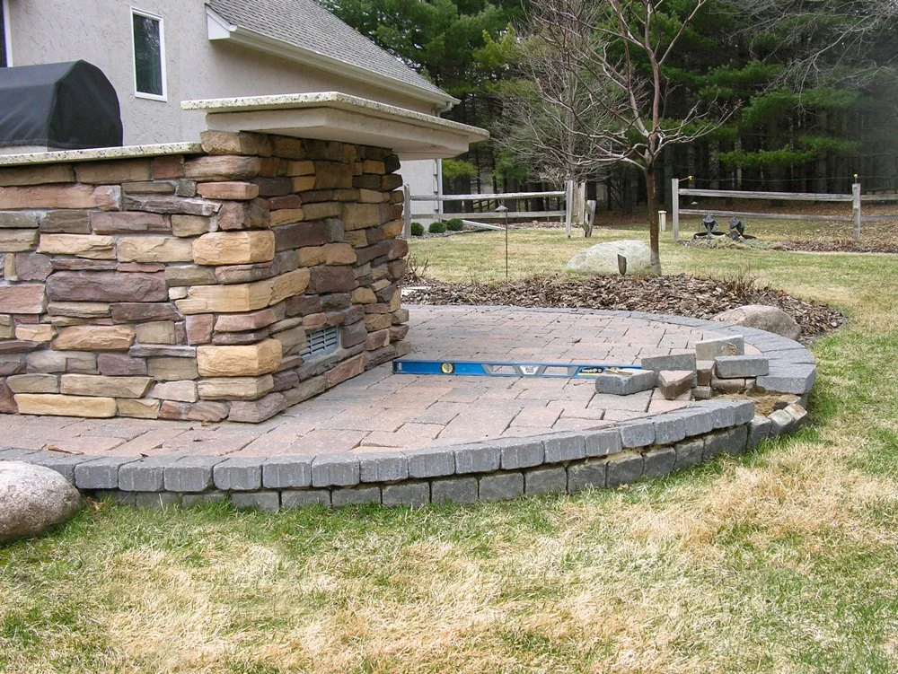 Backyard patio area with stone and brick retaining wall, pavers, and a level, landscaped yard with a tree and a wooden fence in the background.