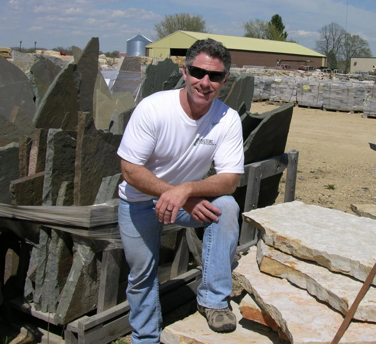 A man smiling and wearing sunglasses, kneeling on a large stone slab at a stone yard with piles of cut stone slabs and a barn in the background.