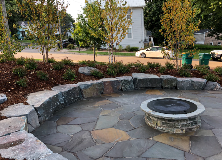 Stone fire pit on a paved patio with a curved stone retaining wall and landscaped yard with trees and bushes