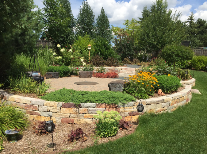 Well-maintained garden with a stone retaining wall, colorful flowering plants, shrubs, trees, and decorative lanterns on a sunny day.