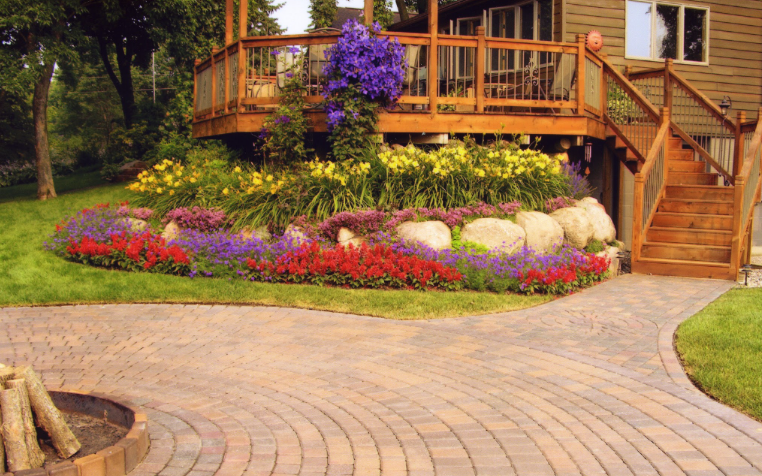 Colorful flower bed with yellow, purple, red, and pink flowers in front of a wooden deck with stairs, a brick-paved pathway, and a house in the background.