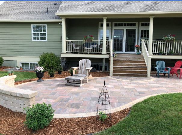 Backyard patio with stone pavers, Adirondack chairs, potted flowers, and a green house with stairs leading to an upper porch.