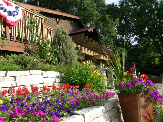 Garden with colorful flowers, potted plants, and a wooden house with a deck in the background, surrounded by greenery.