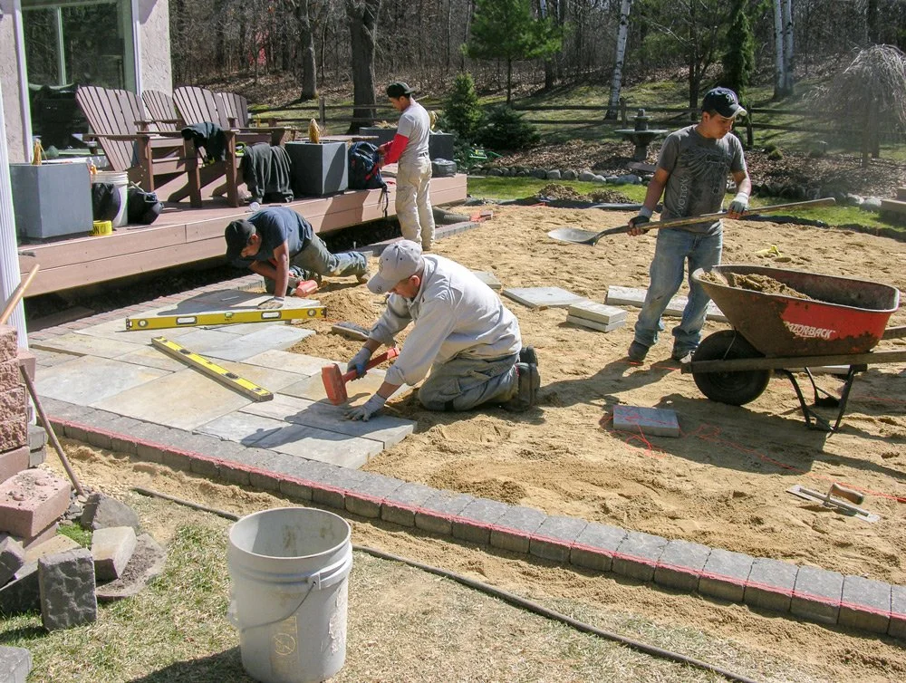 People working on laying stone pavers in a backyard landscaping project, with some workers kneeling and others standing, using equipment like a level and a wheelbarrow.
