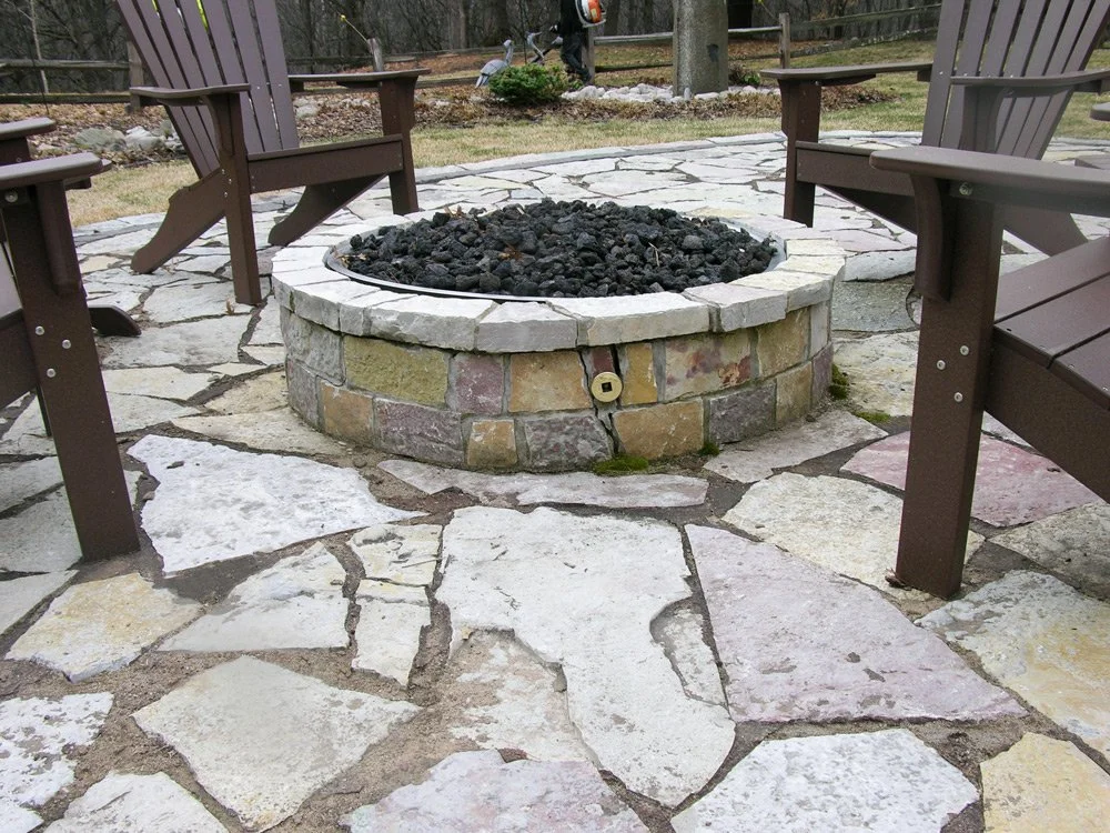Fire pit with black rocks surrounded by four wooden chairs on a stone patio in an outdoor setting.