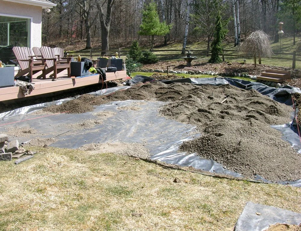 Backyard with a patio, chairs, and a garden bed under construction, with piles of dirt and landscaping materials.