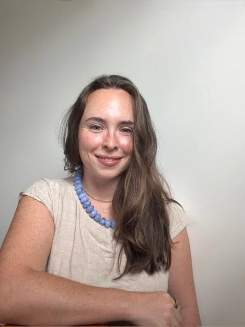 A young woman with long brown hair smiling at the camera, wearing a beige top and a blue beaded necklace, sitting in front of a plain light-colored wall.