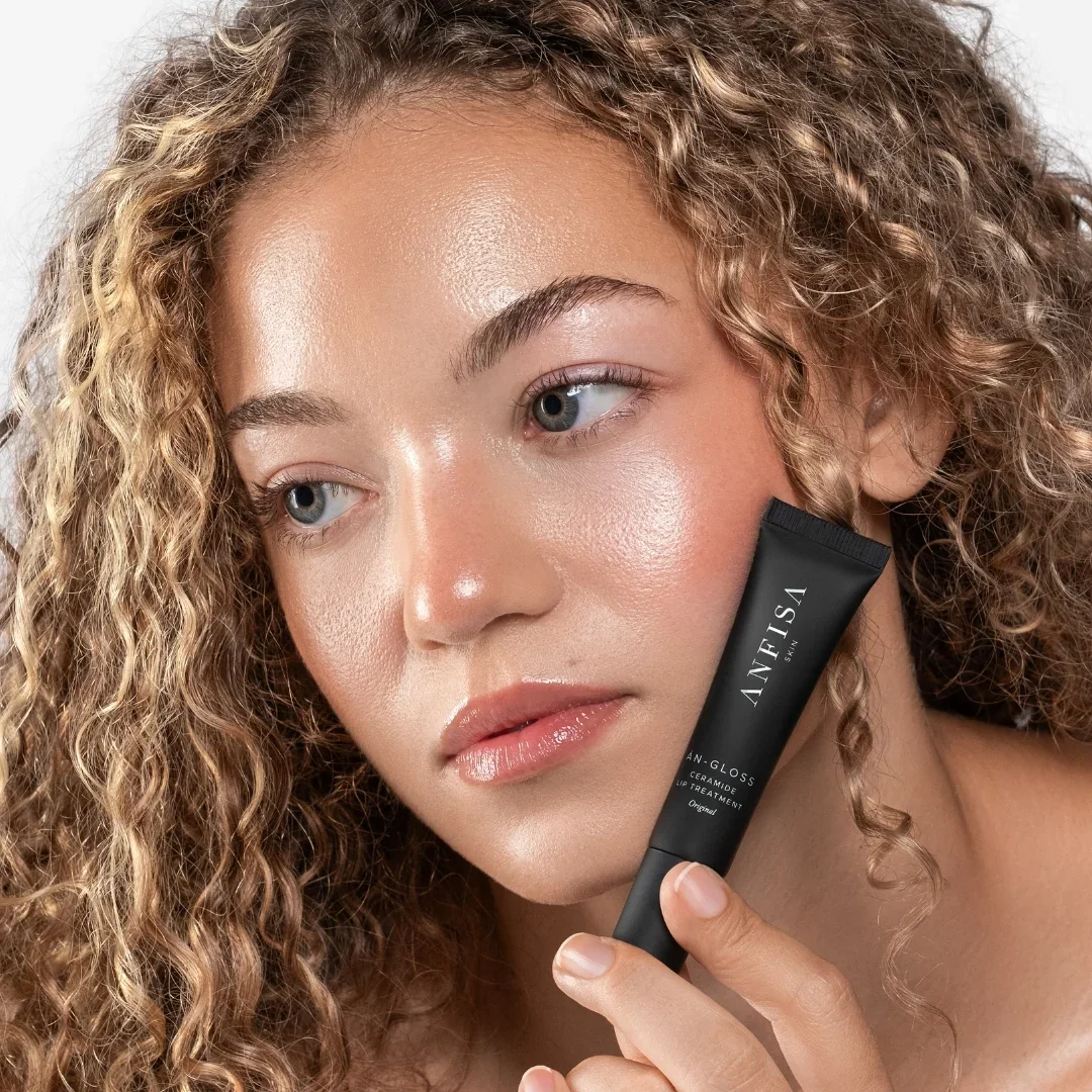 A young woman with curly blonde hair holding a black tube of Anfisa skincare product near her face