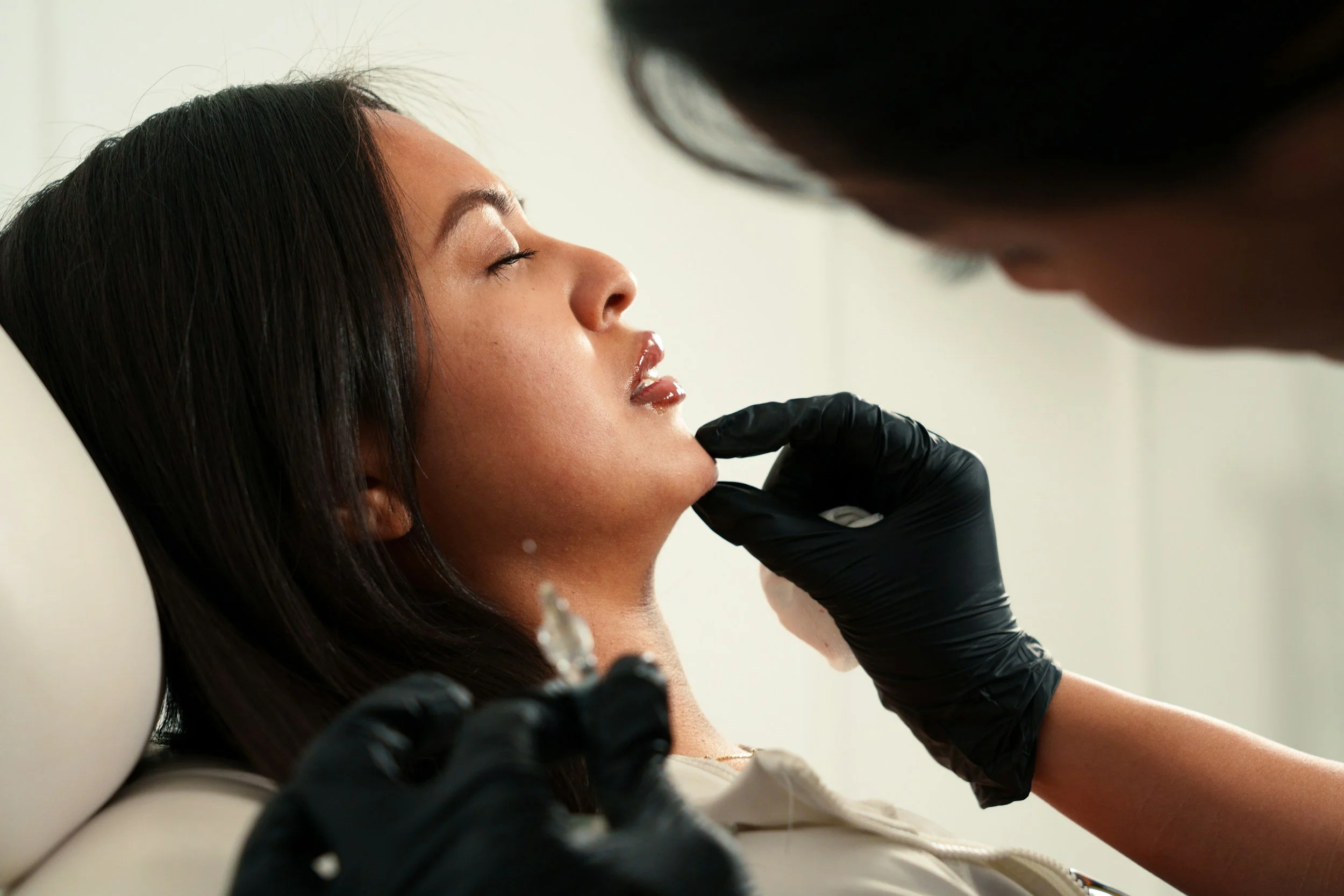 A woman lying back with her eyes closed as a person in black gloves examines her face and prepares to inject dermal filler in her chin.