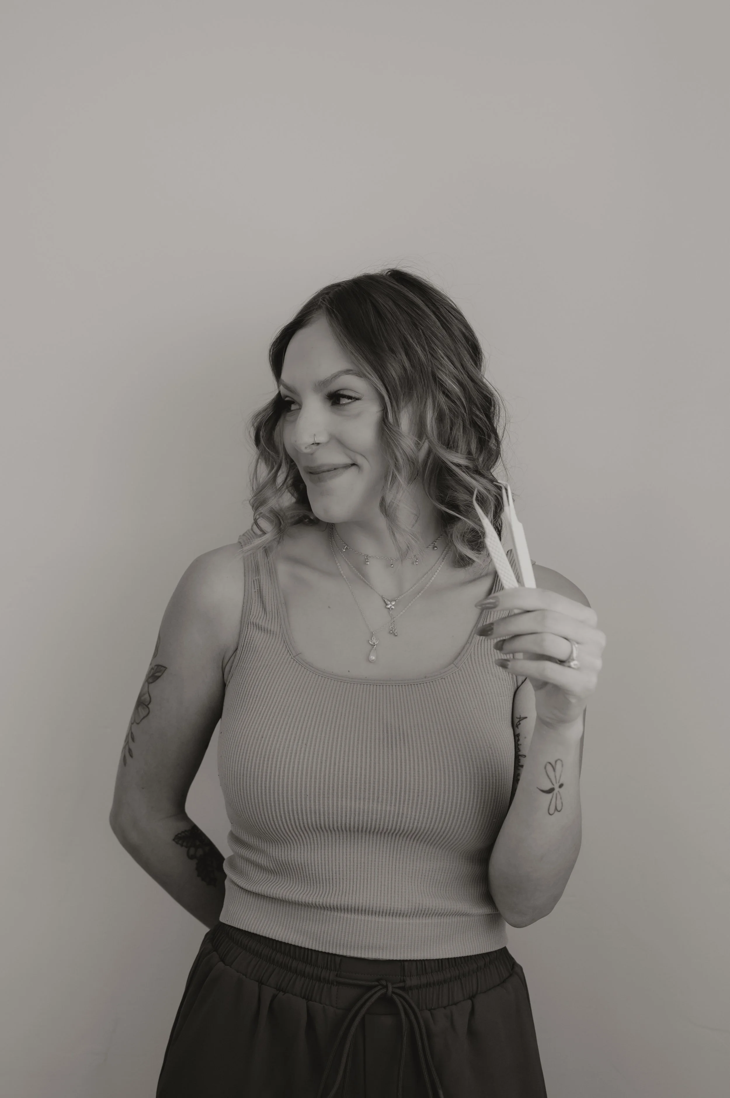 A woman with wavy hair, wearing a tank top and patterned pants, holding a straw and smiling while looking to her left against a plain background.