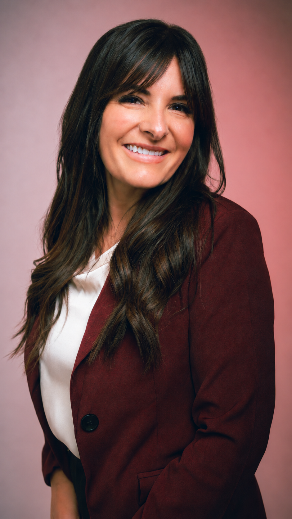 A woman with long brown hair, wearing a white top and a maroon blazer, smiling against a pink gradient background.