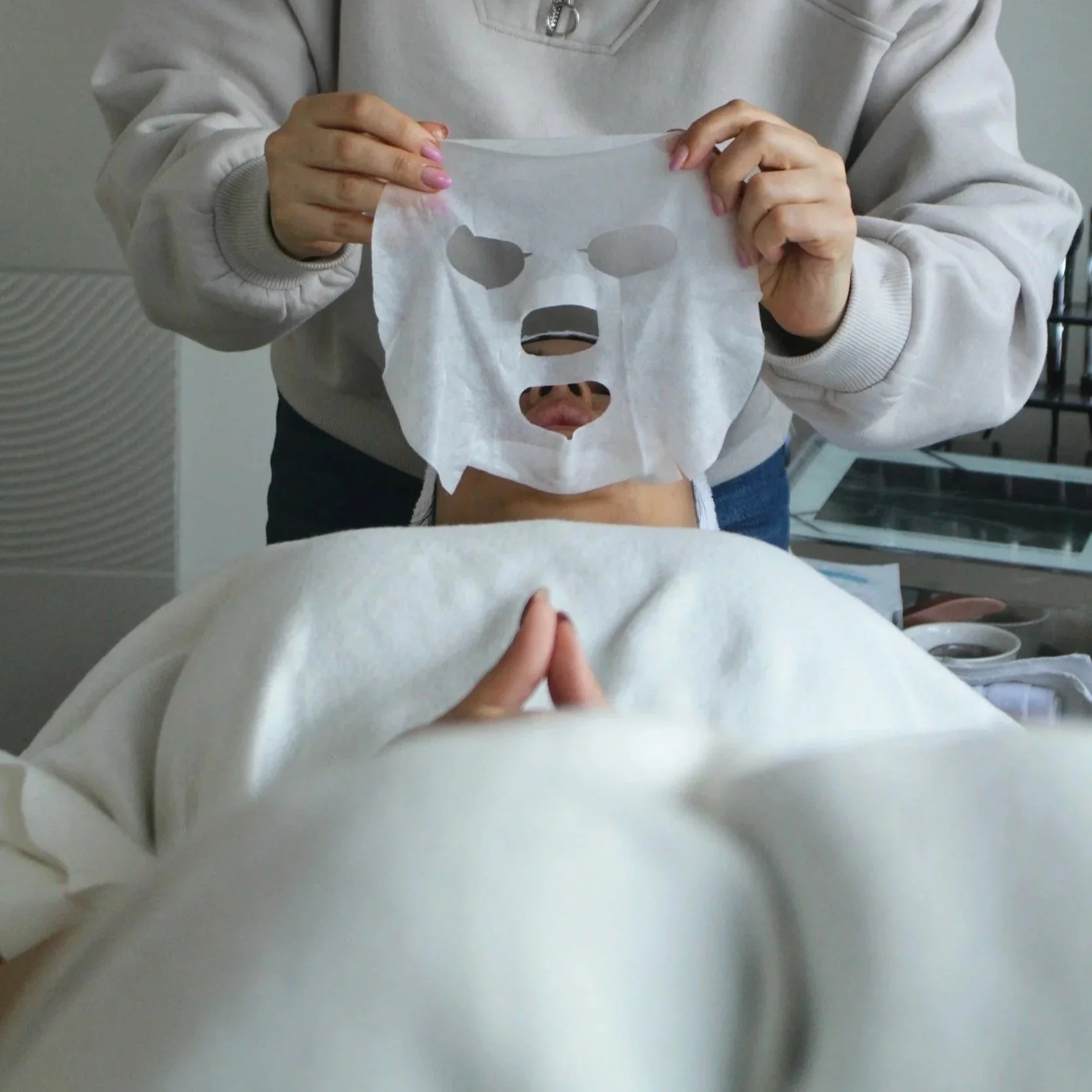Person laying on a facial treatment bed with a luxury skincare sheet mask above their face, with another person in a beige sweater adjusting it.